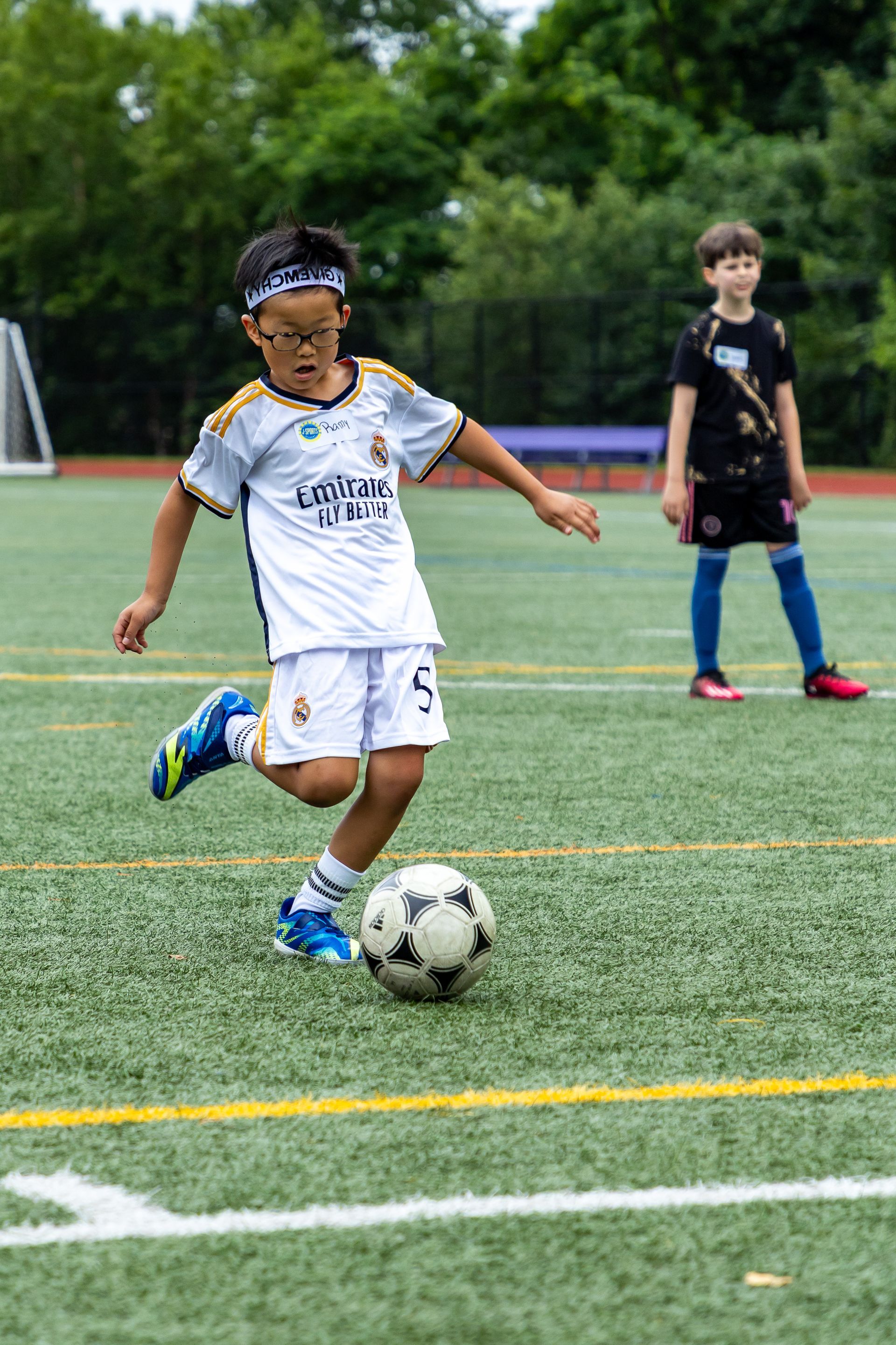 Boy in soccer uniform kicks a soccer ball on a green field, another boy in the background.