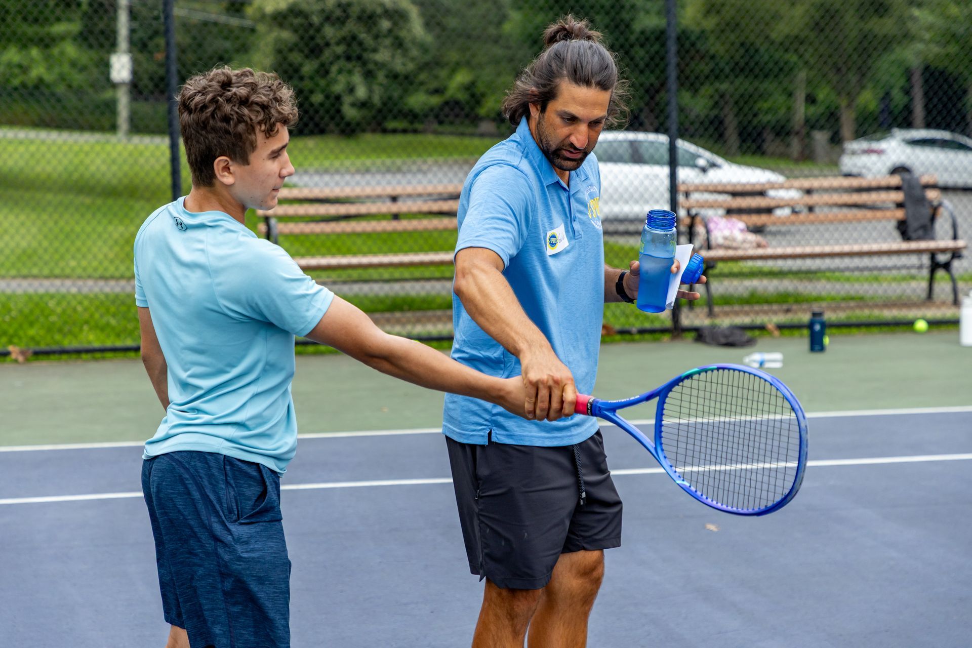 A tennis coach instructs a young player on a court. The coach, in a blue shirt, holds the racket and guides the boy's grip.
