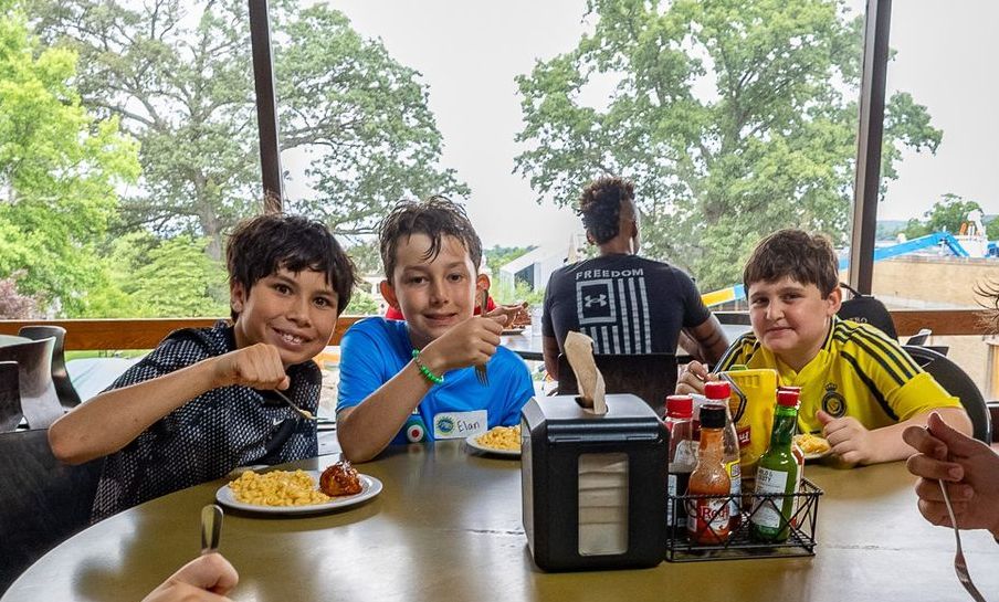 Three boys smiling and pointing at the camera while eating at a table, with a person in the background.