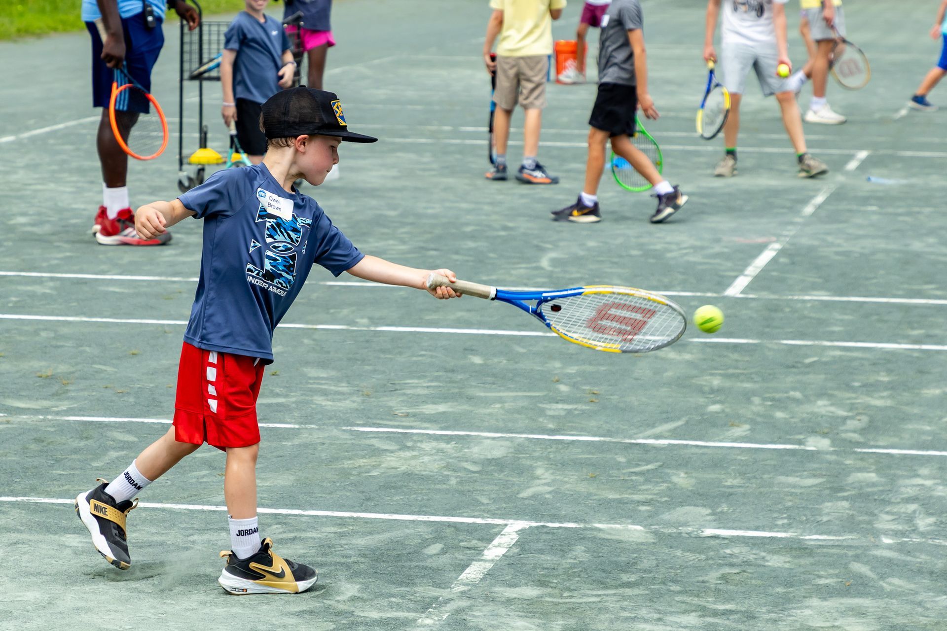 Boy in red shorts swings tennis racket at a ball on an outdoor court with other children in background.