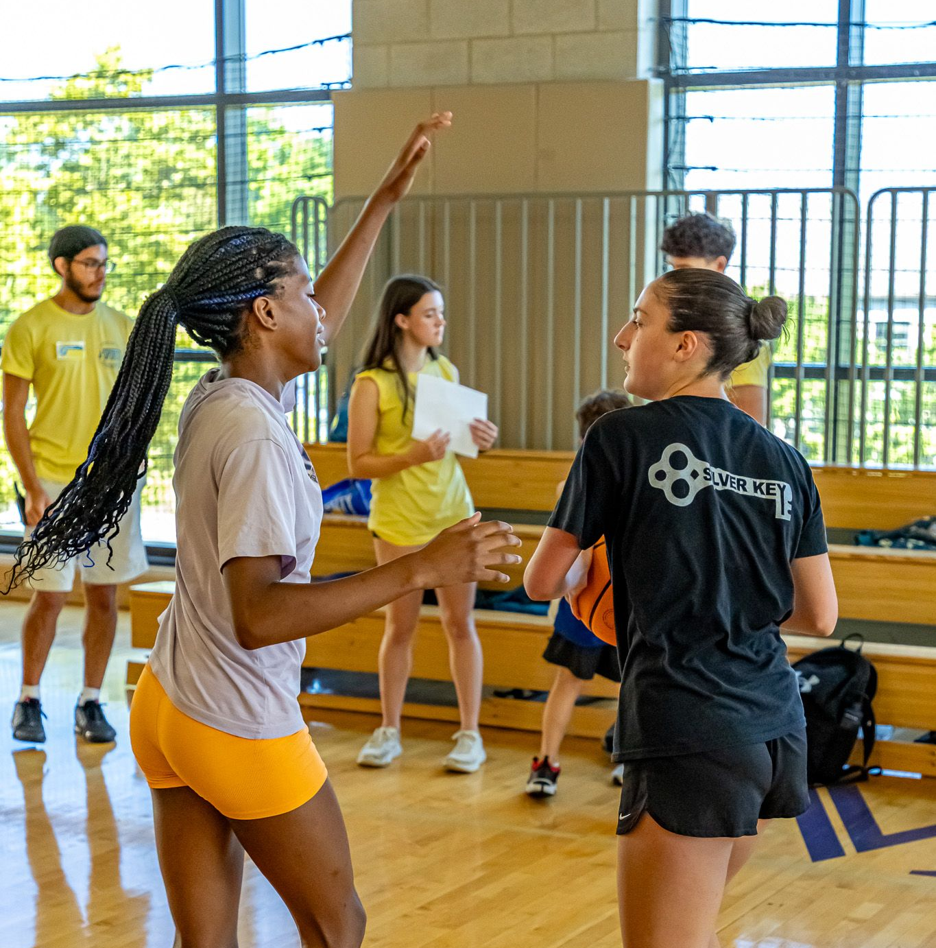 Basketball game: Players indoors, one with raised arm about to throw, others watching in a gym.