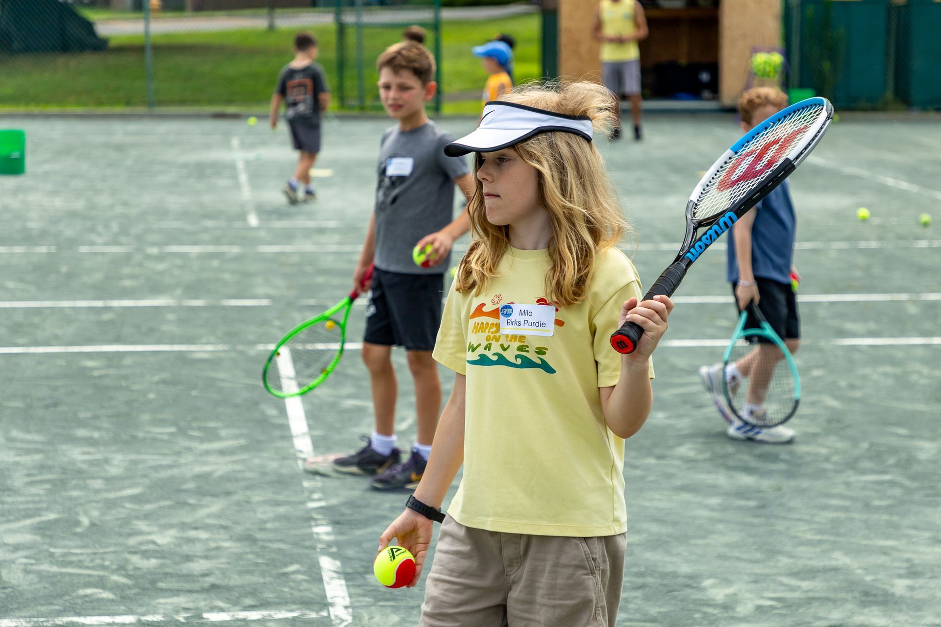 Boy with curly hair on tennis court, holding a racket and a ball, wearing a visor.