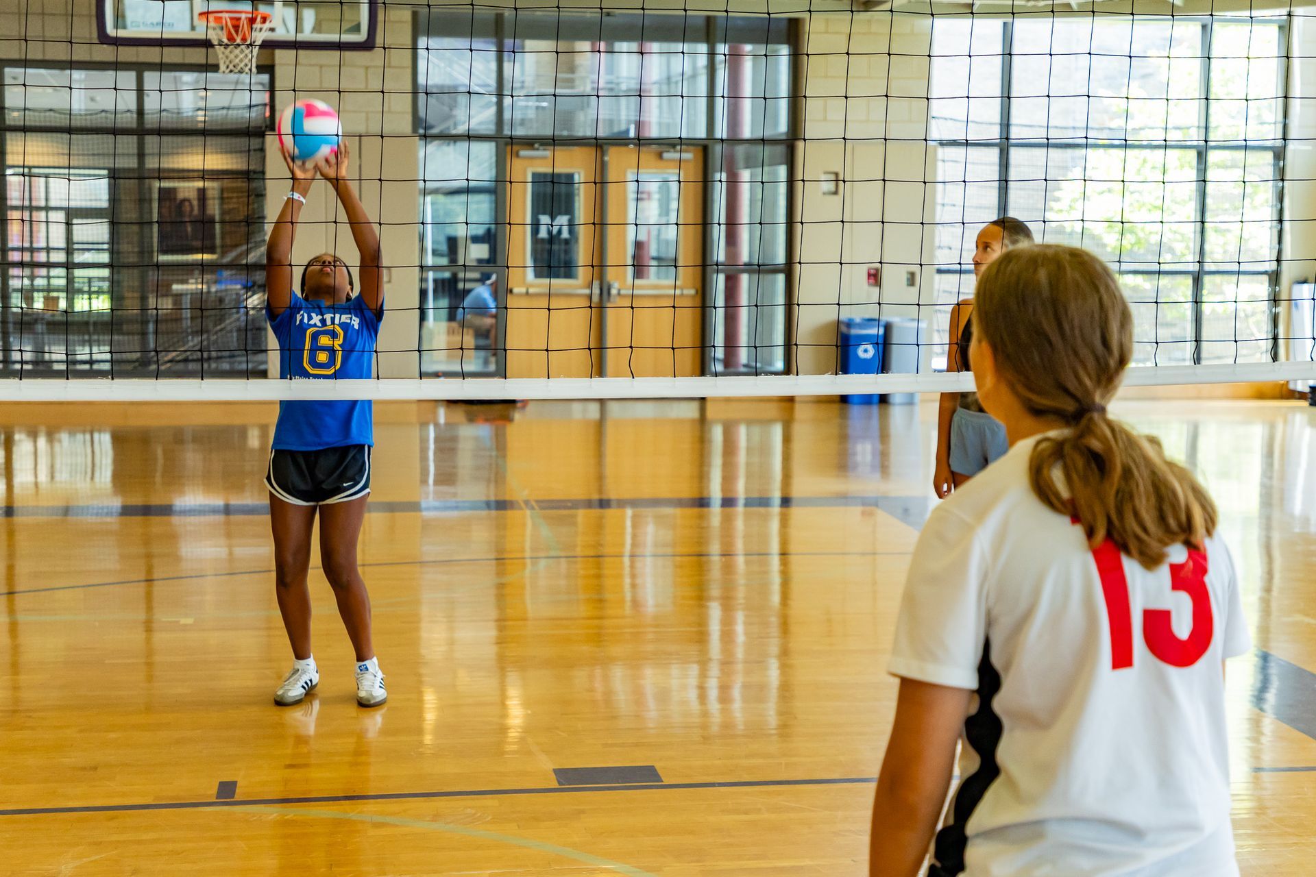 Volleyball player in blue uniform sets the ball, another player in white shirt watches in a gym.
