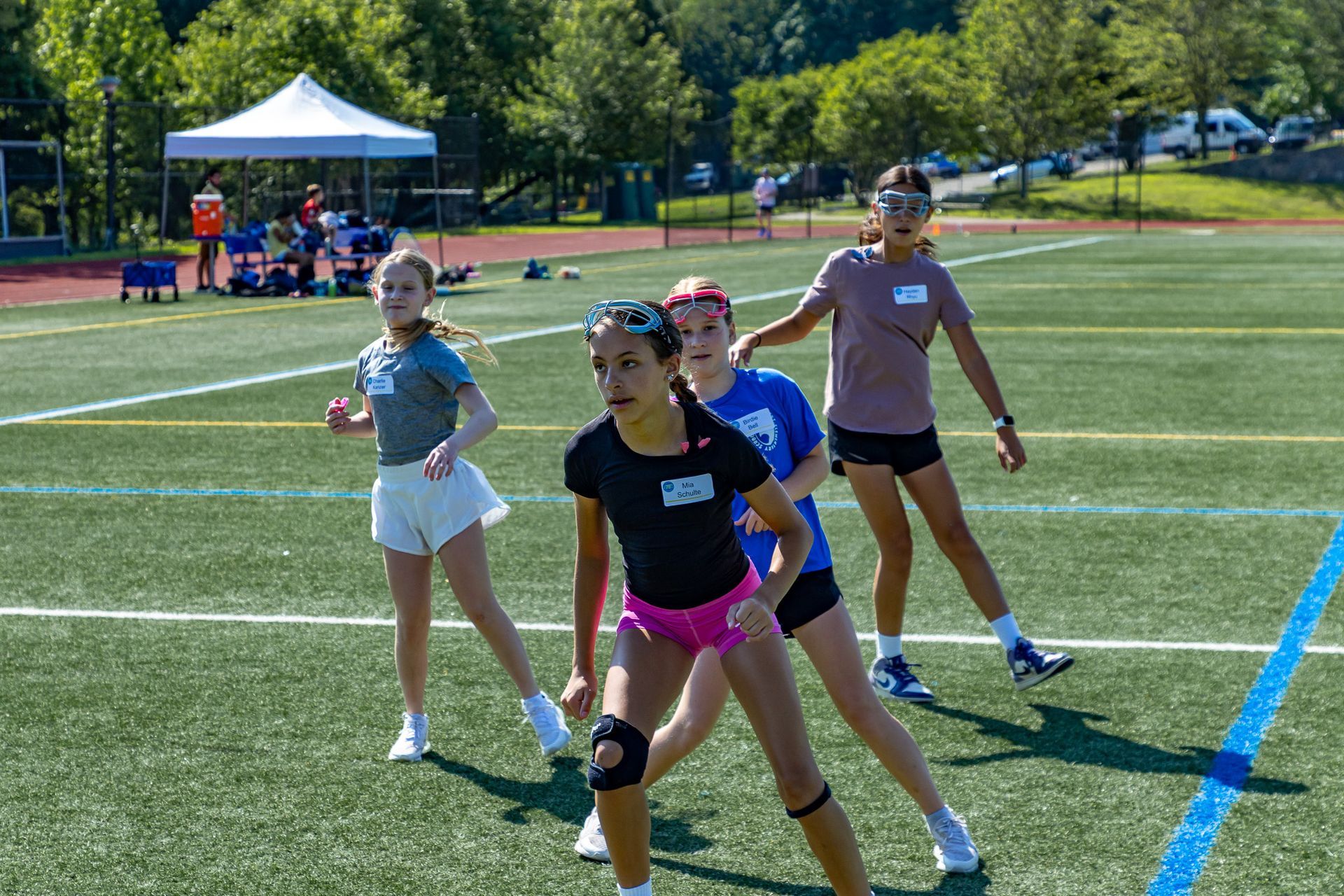 Girls in athletic gear on a green field, some stretching, under a sunny sky.