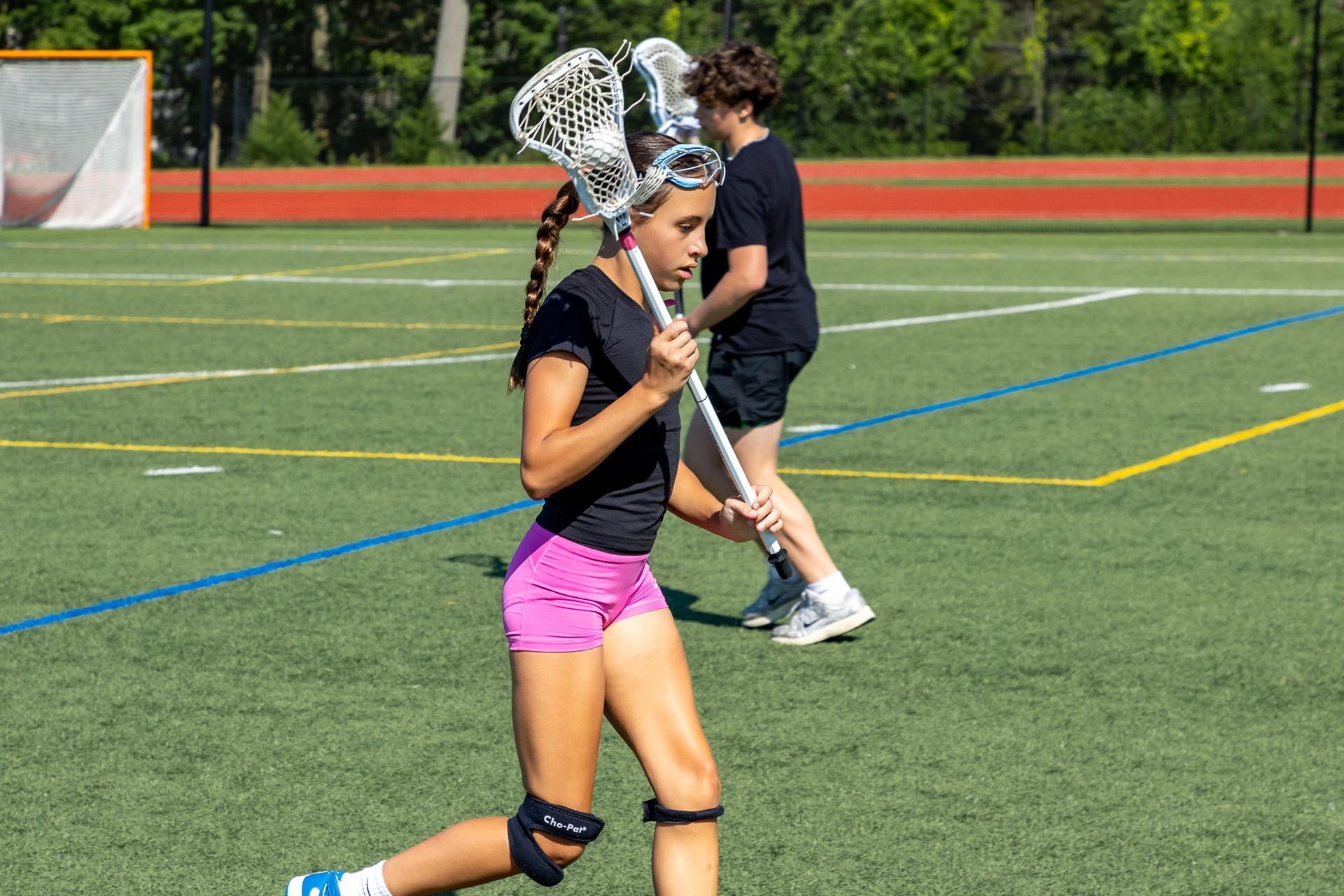 Girl in pink shorts and black shirt with lacrosse stick on green field.