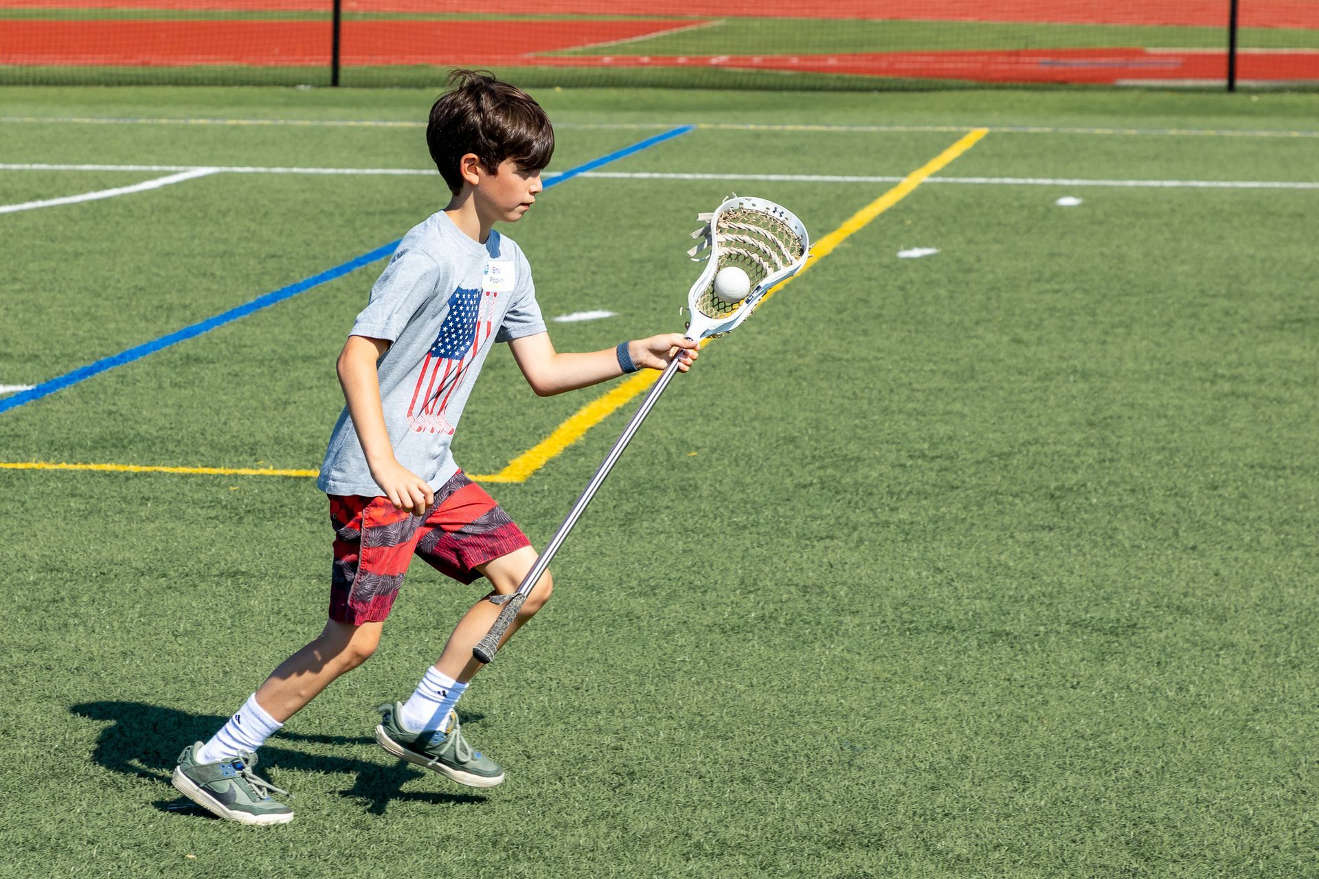 Boy playing lacrosse on a green field, wearing a patriotic shirt, holding the ball in his lacrosse stick.