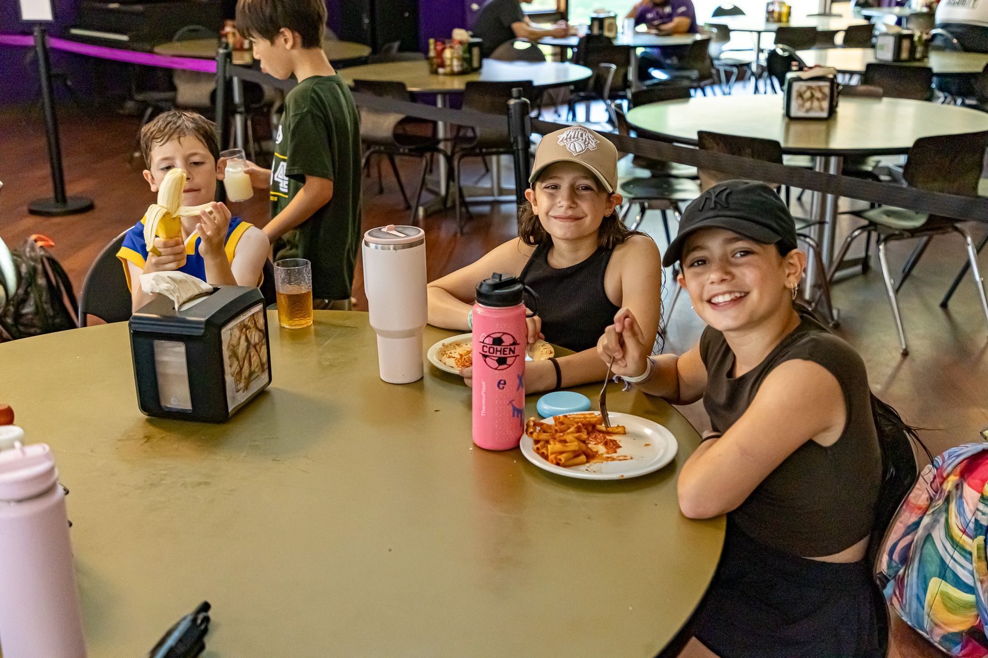 Three kids smile while eating at a table in a cafeteria. One eats pasta, another a banana.