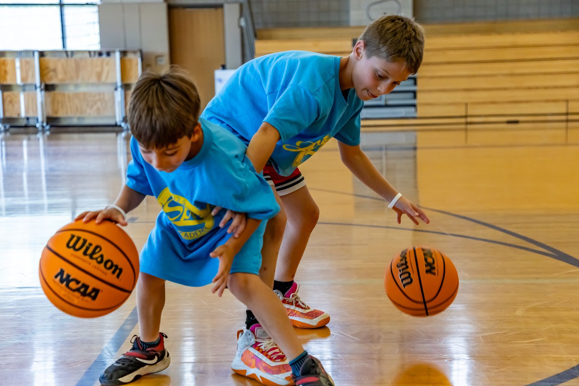 Two boys in blue shirts dribble basketballs on a wooden court.