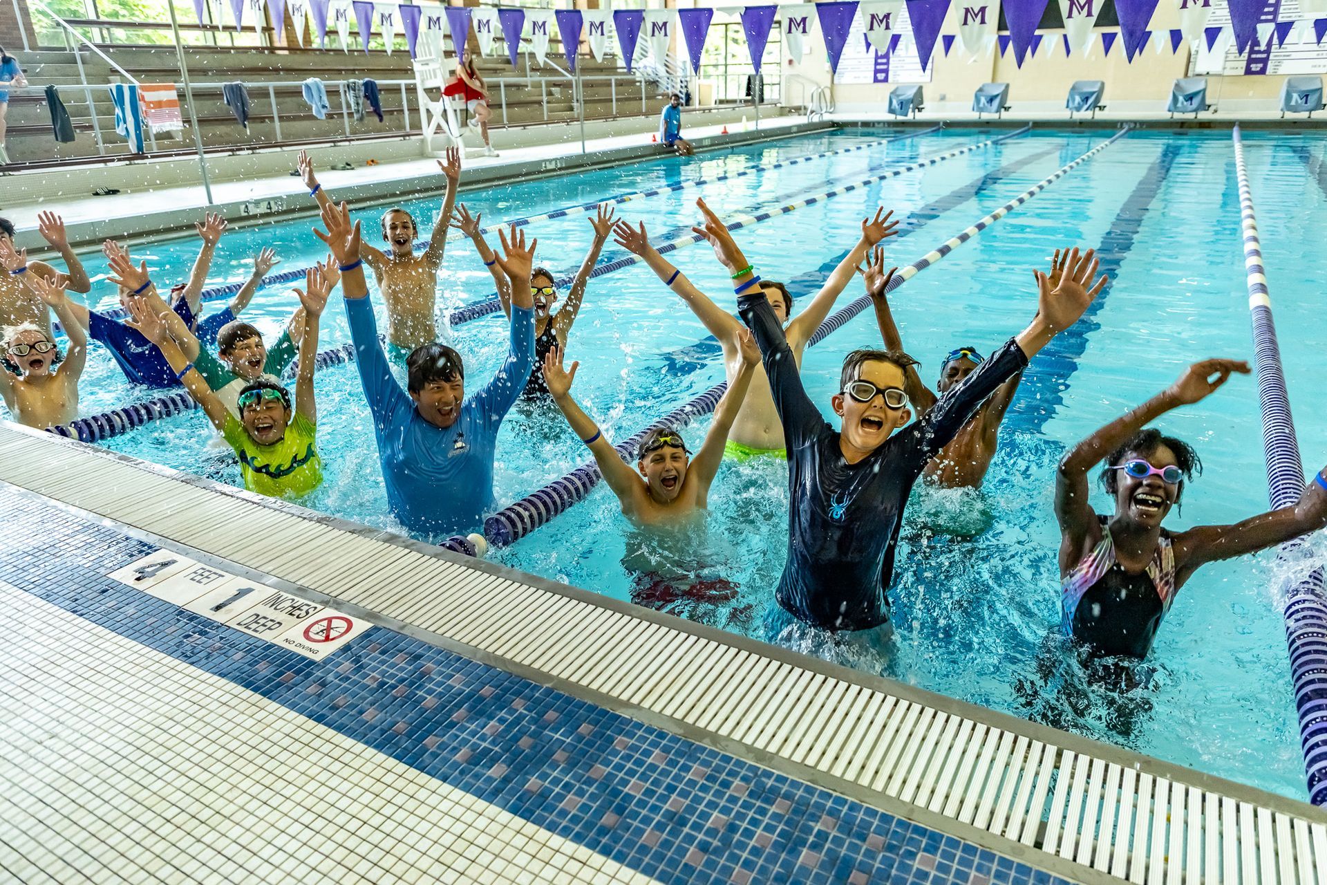 Group of children in a pool cheering, arms raised, with smiles, blue water and lanes.