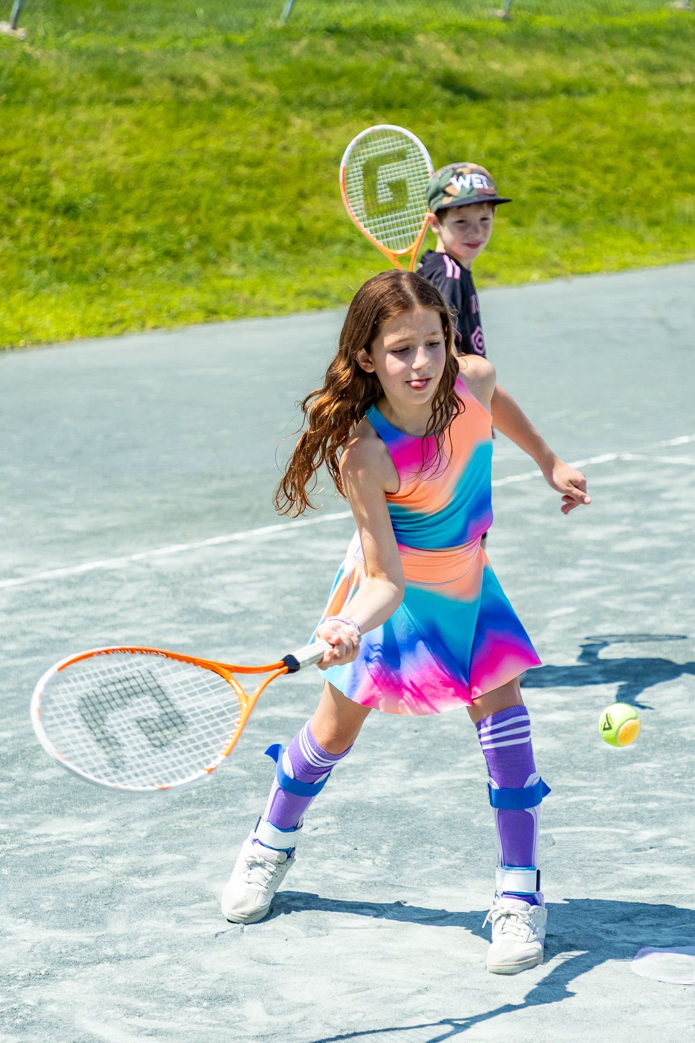 Girl in colorful tennis outfit swings at a ball on a court, boy behind.