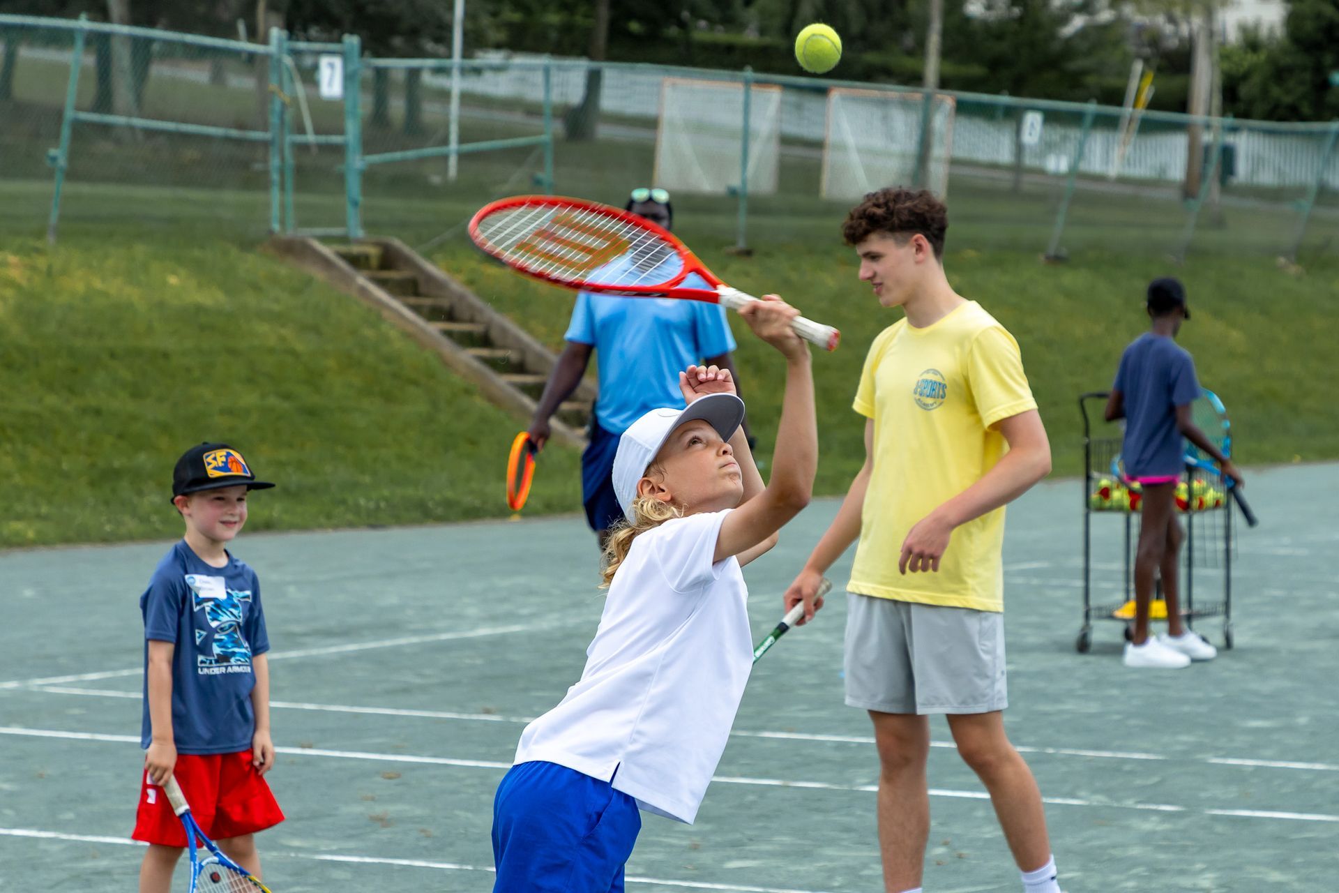Boy swinging tennis racket with coach on court; other kids watch.
