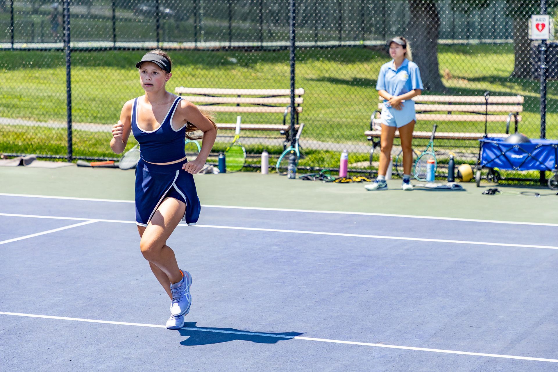 Woman in navy tennis outfit runs on a blue court; another woman watches in the background.