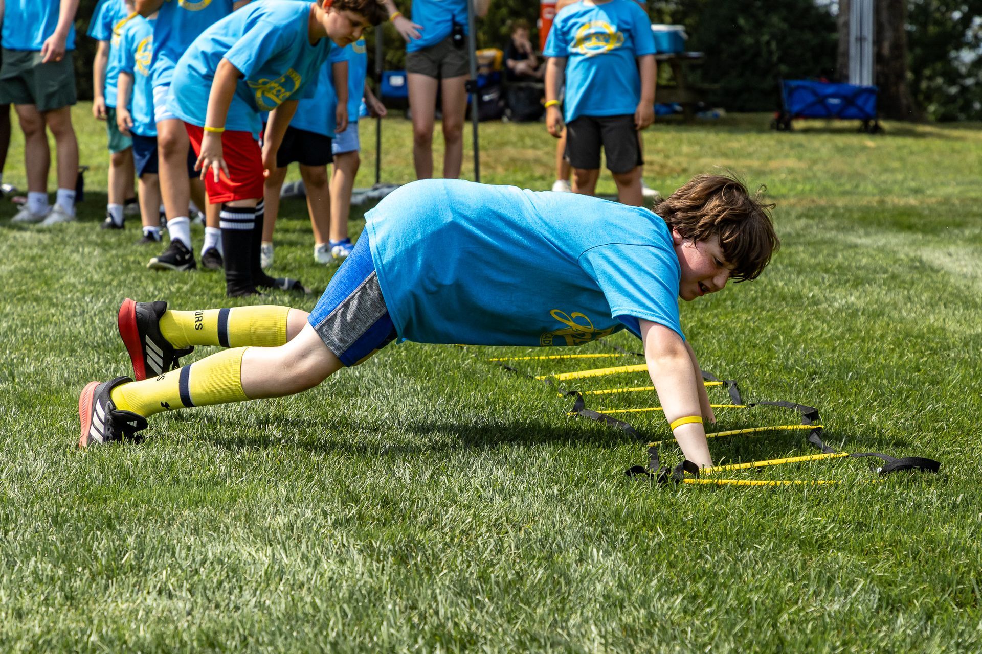 Boy doing push-ups on a ladder on a grass field, wearing a blue shirt with other kids in the background.