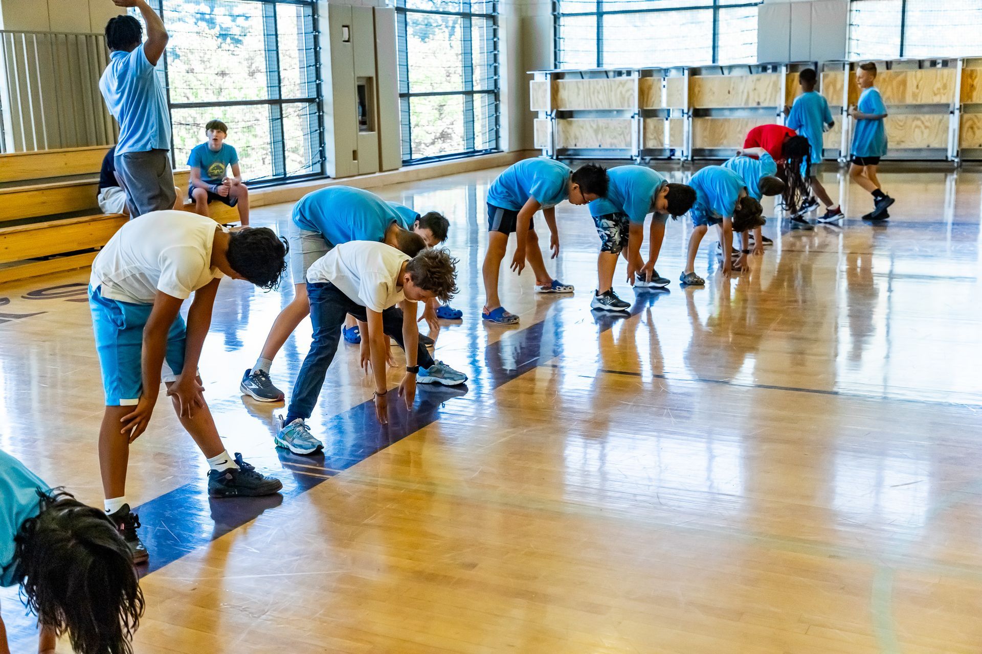 Children in blue shirts stretching on a wooden gym floor.
