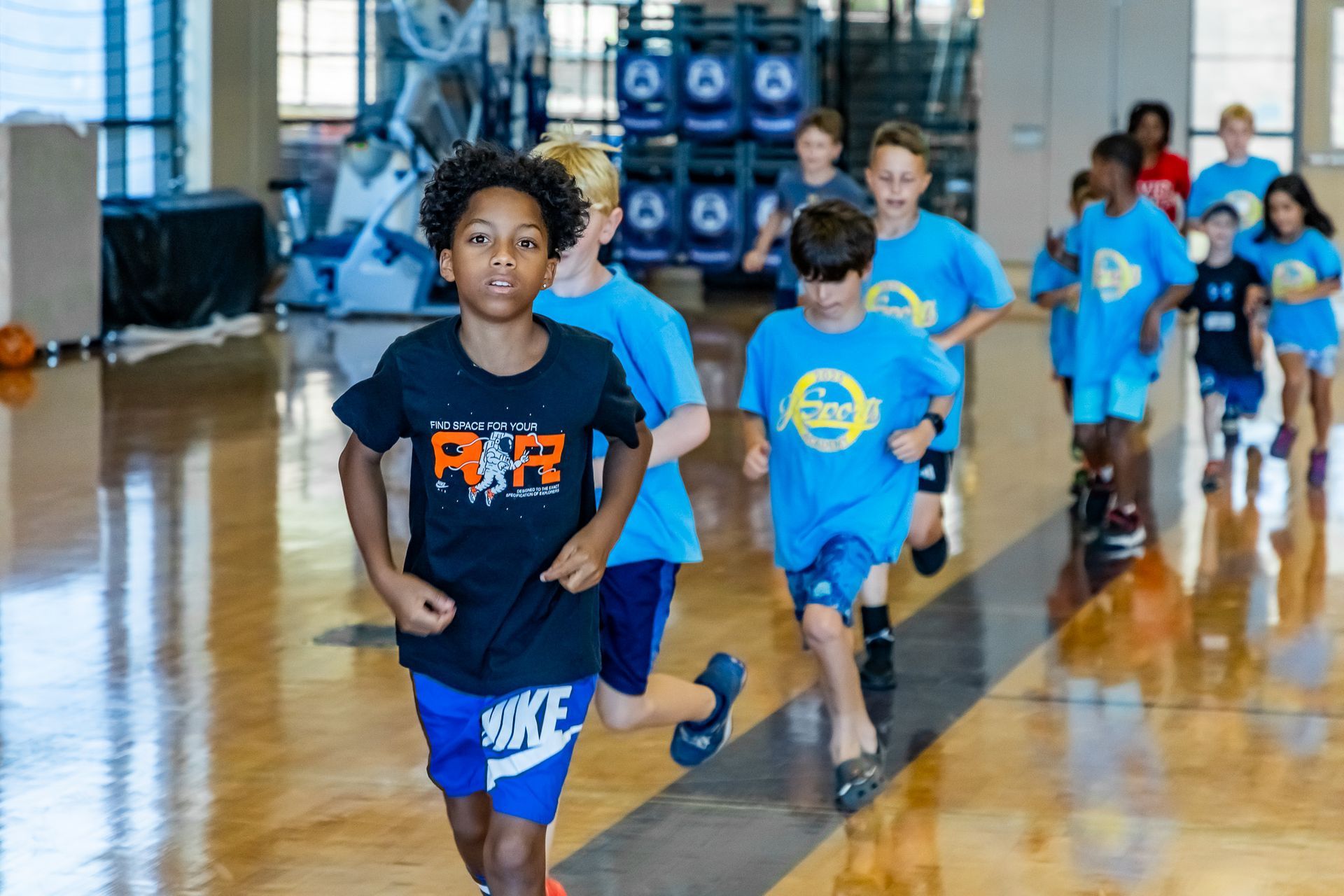 Boys in athletic wear running on a wooden floor indoors, with a few wearing blue t-shirts with a logo.