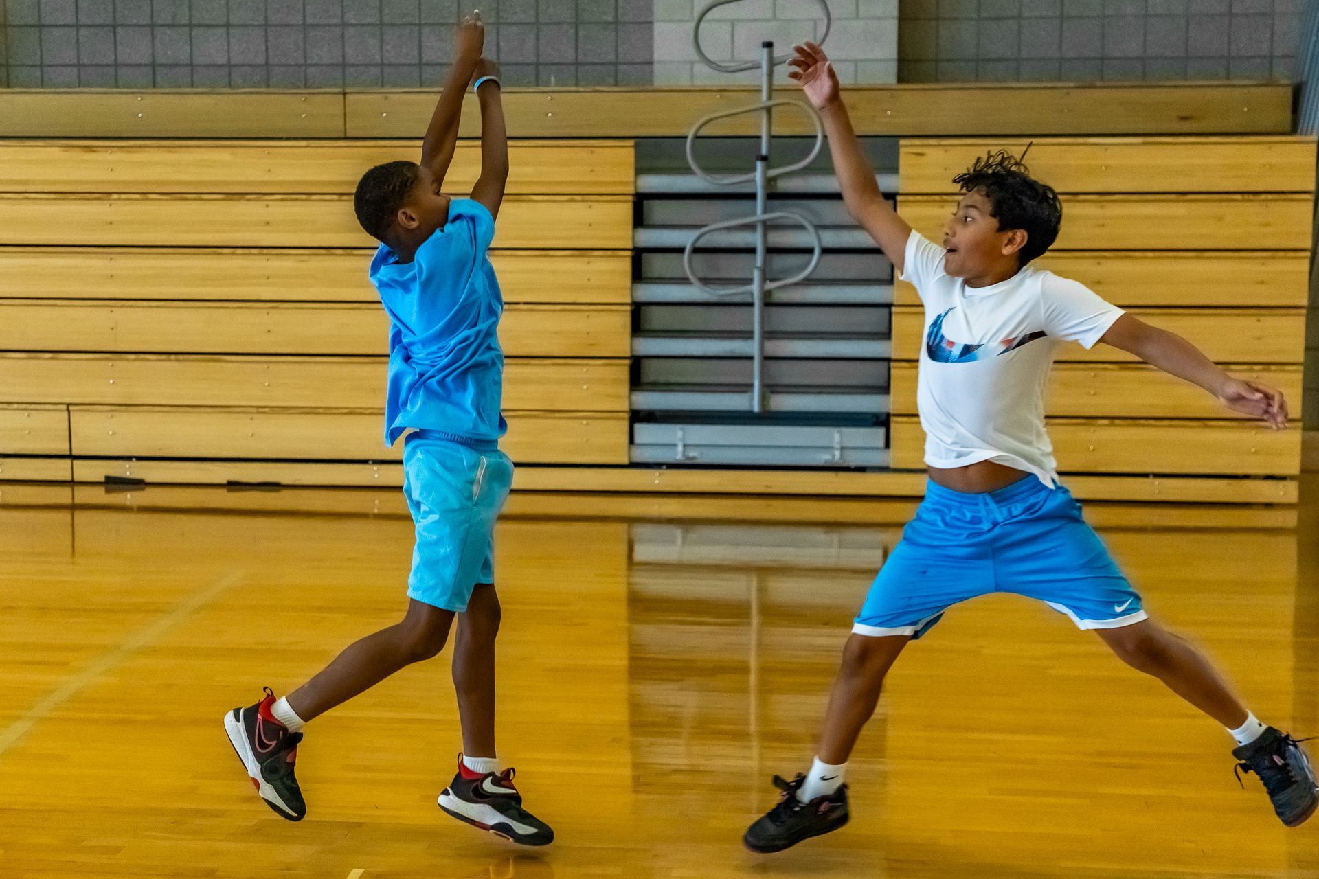 Two boys playing basketball in a gym, one shooting with the other defending.