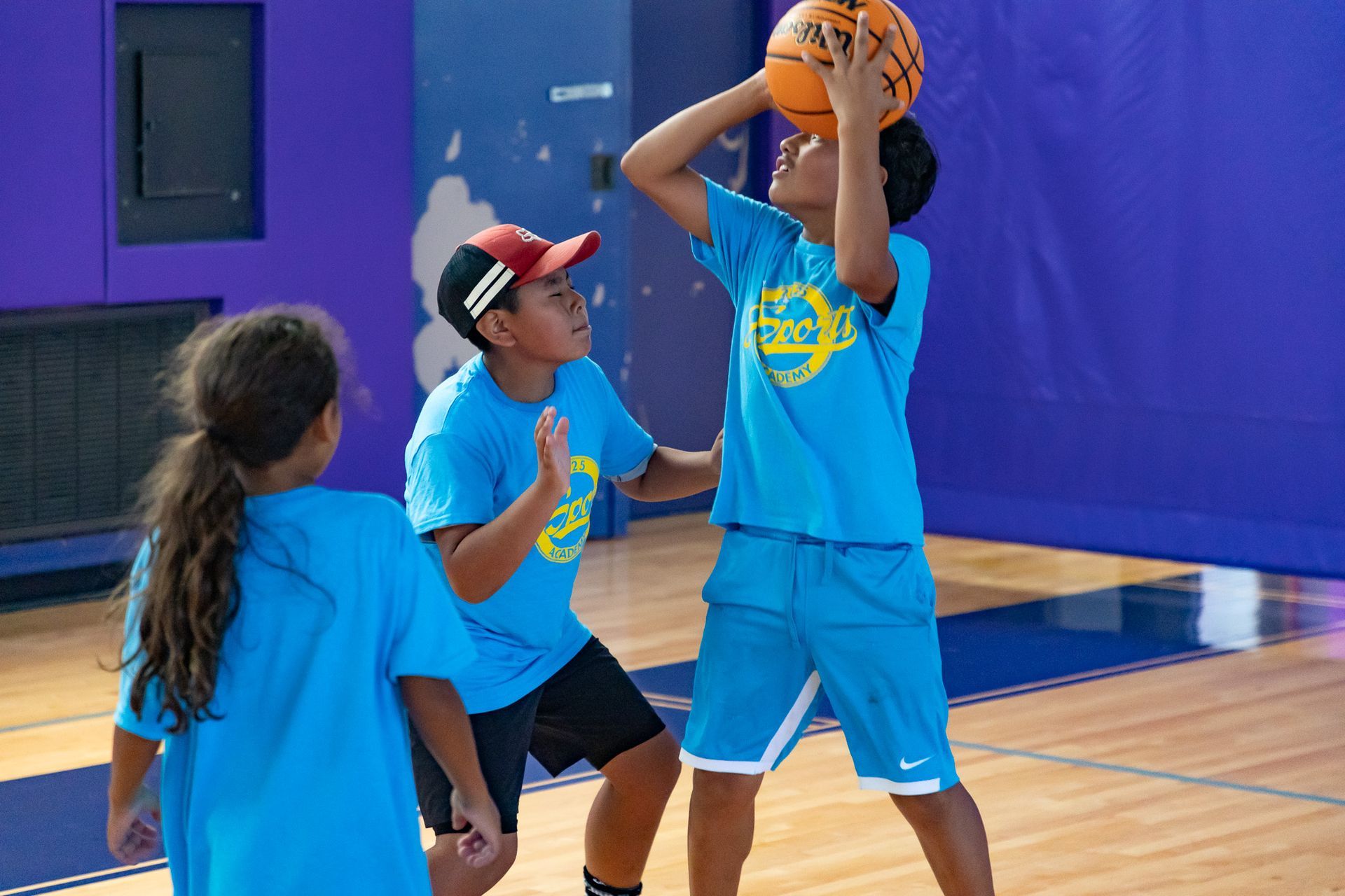 Kids playing basketball in a gym. One shooting, two watching. Wearing blue shirts.
