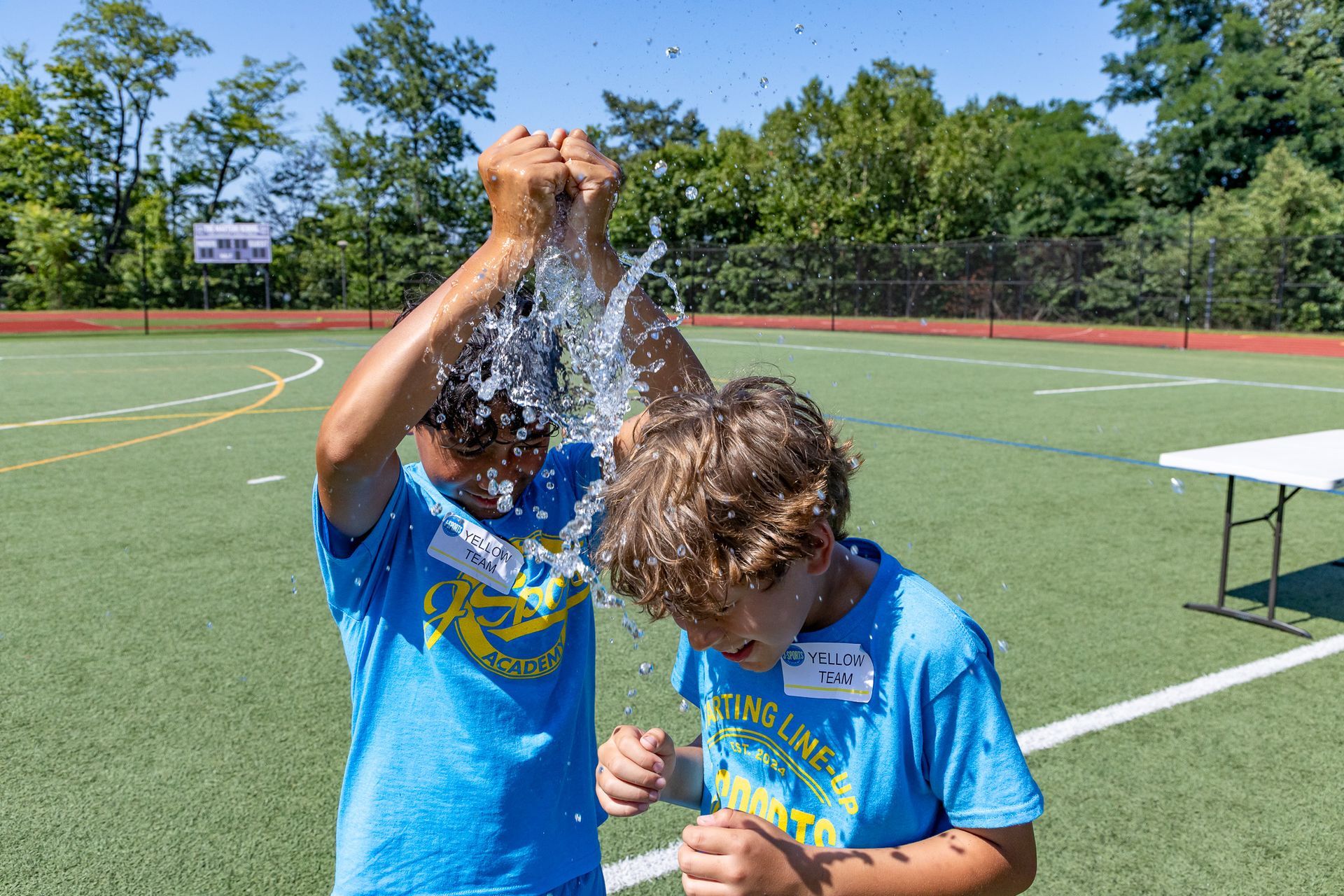 Two boys in blue shirts on a sports field are splashed with water on a sunny day.