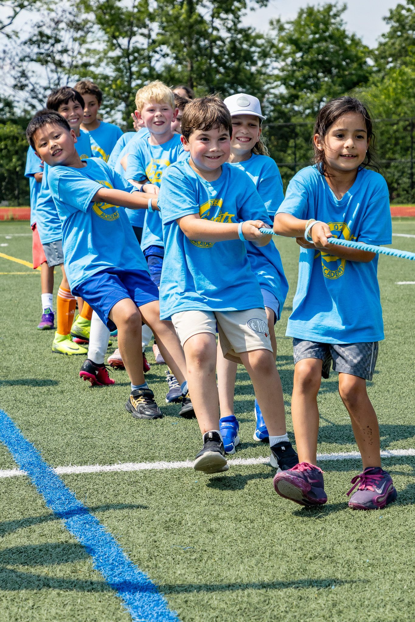 Children in blue shirts playing tug-of-war on a green field, smiling and focused.