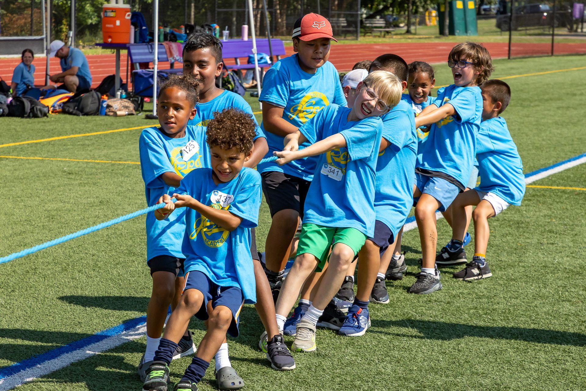 Children in blue shirts playing tug-of-war on a green field, sunny day.