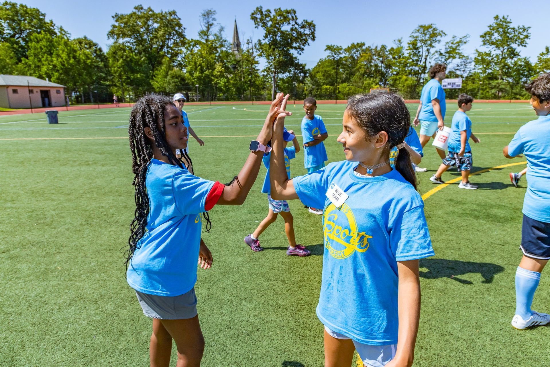 Two girls in blue shirts high-five on a green field during a sunny day camp activity. Other children and trees are visible.