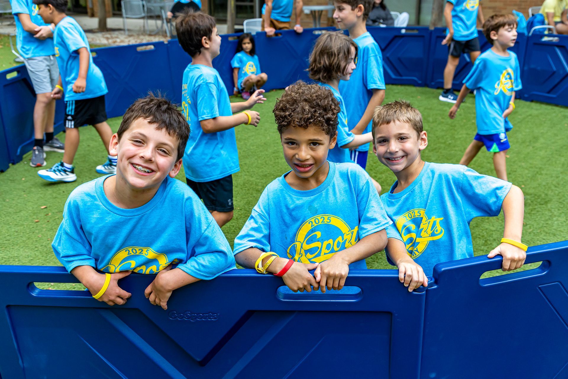 Smiling children in blue shirts and wristbands, in an enclosed play area.