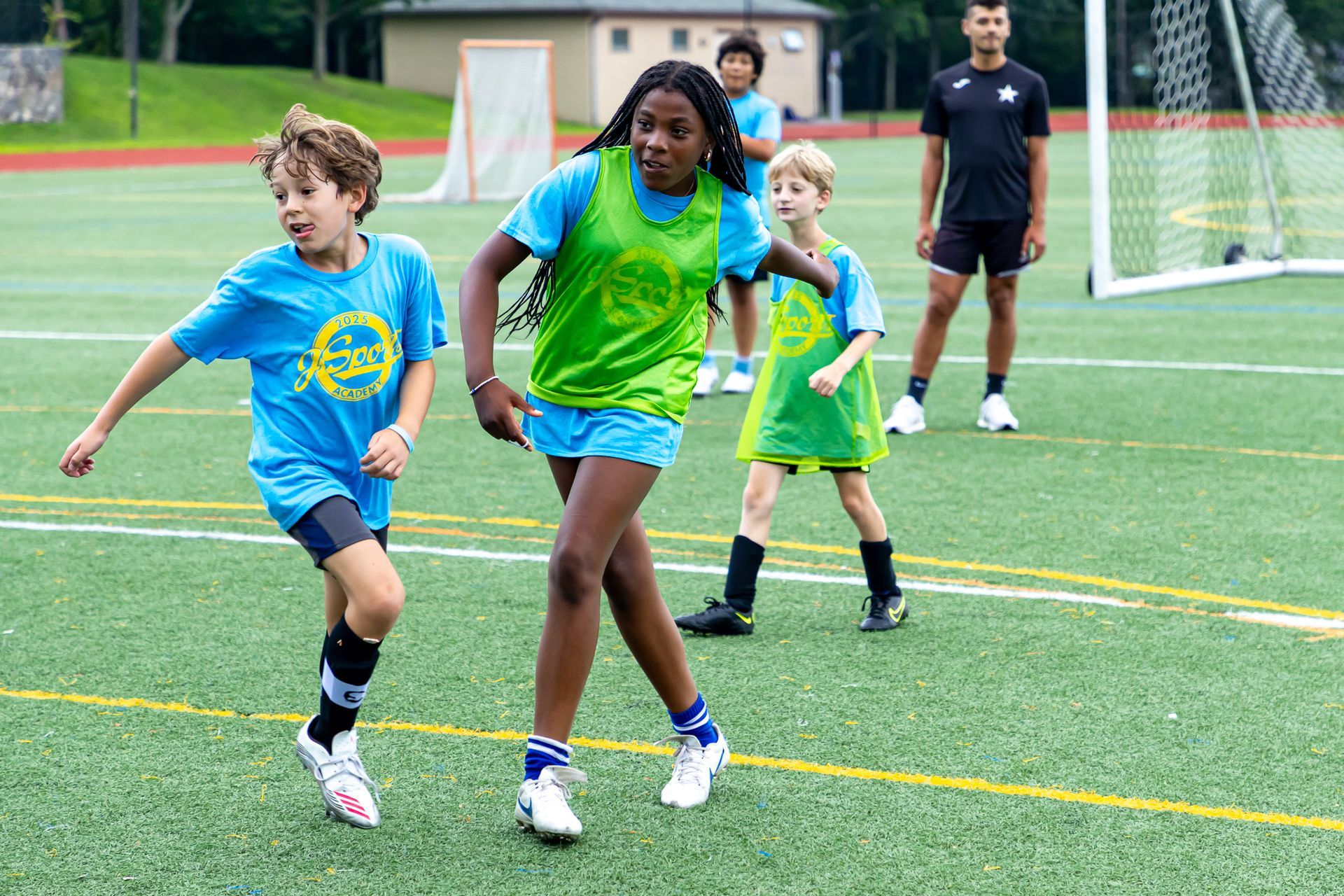 Children in blue shirts play soccer on a green field, with a coach observing.