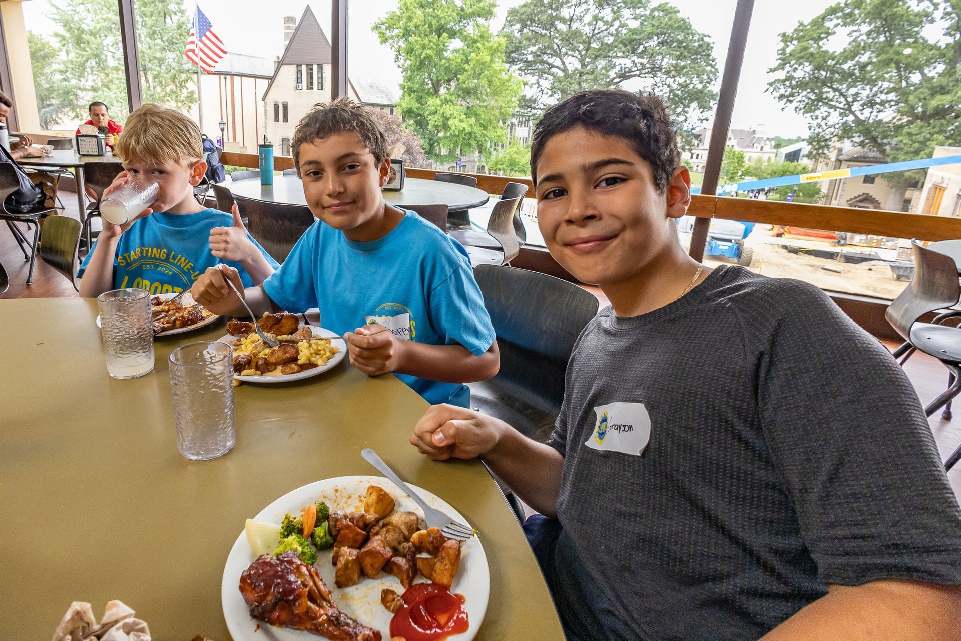 Three boys smiling and eating a meal at a table, indoors with an American flag in the background.