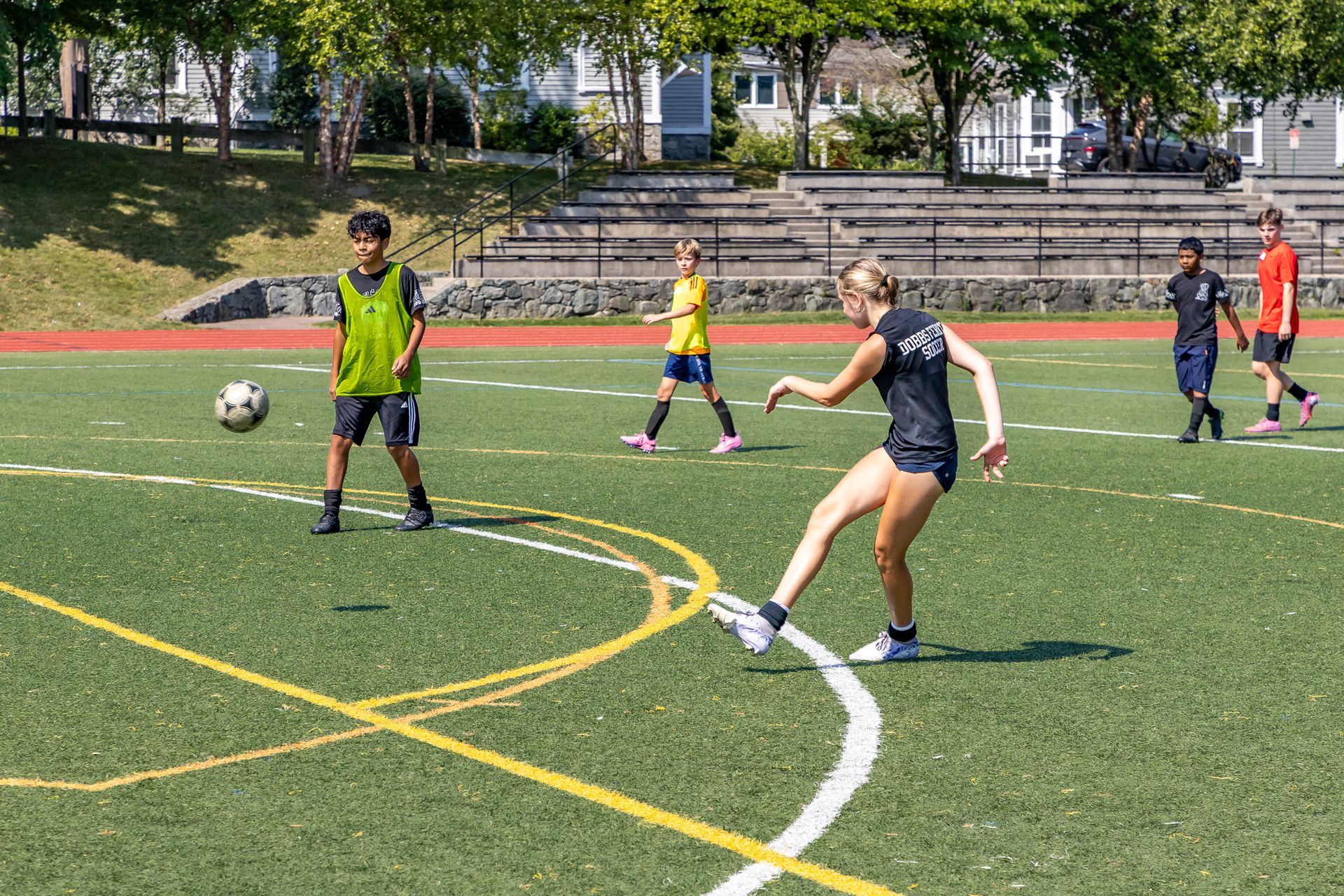 Soccer players on a green field; a woman kicks the ball, a boy in a vest stands ready.