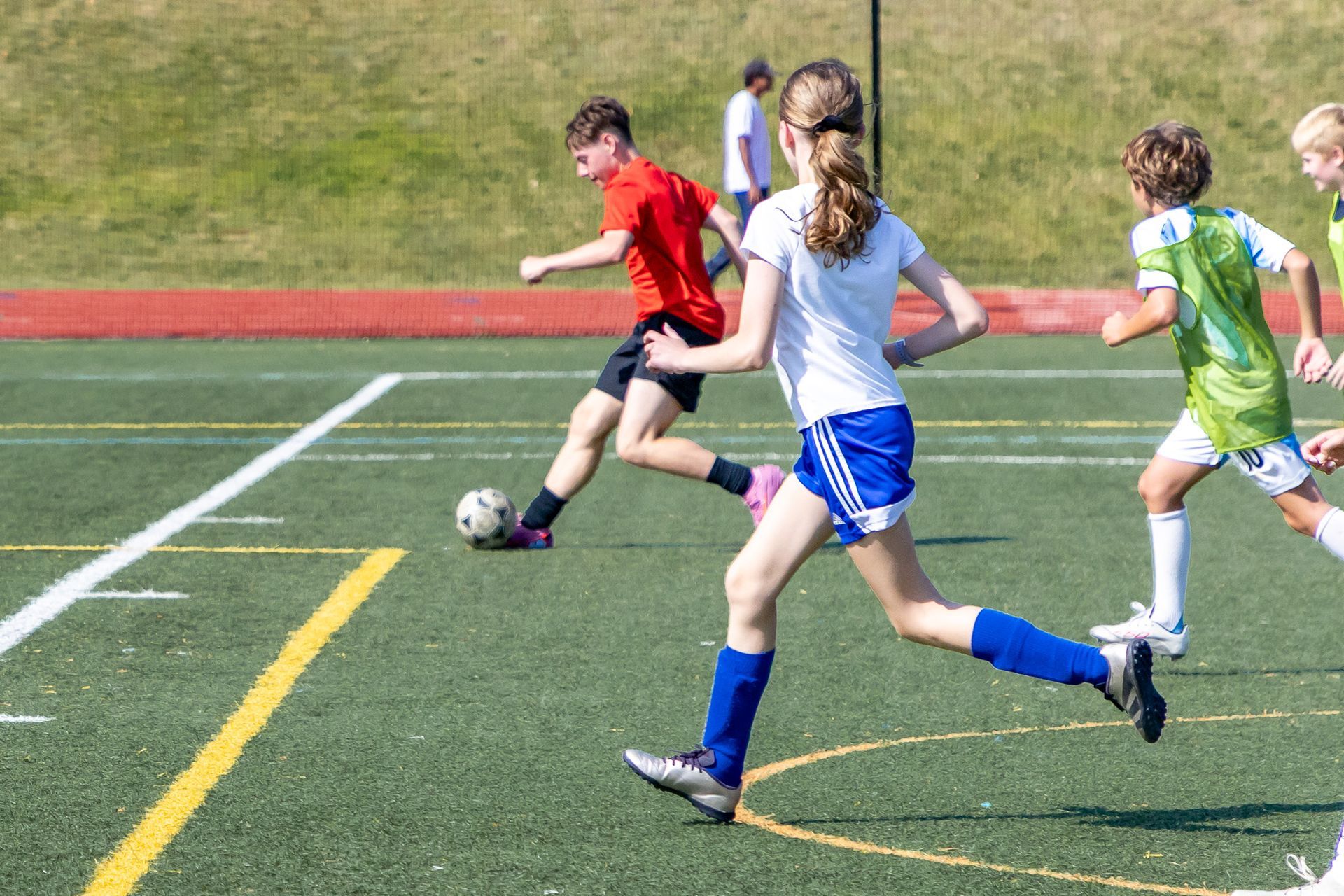 Soccer game: Teenagers on a green field, running and kicking the ball. One in red shirt, others in blue, green, and white.