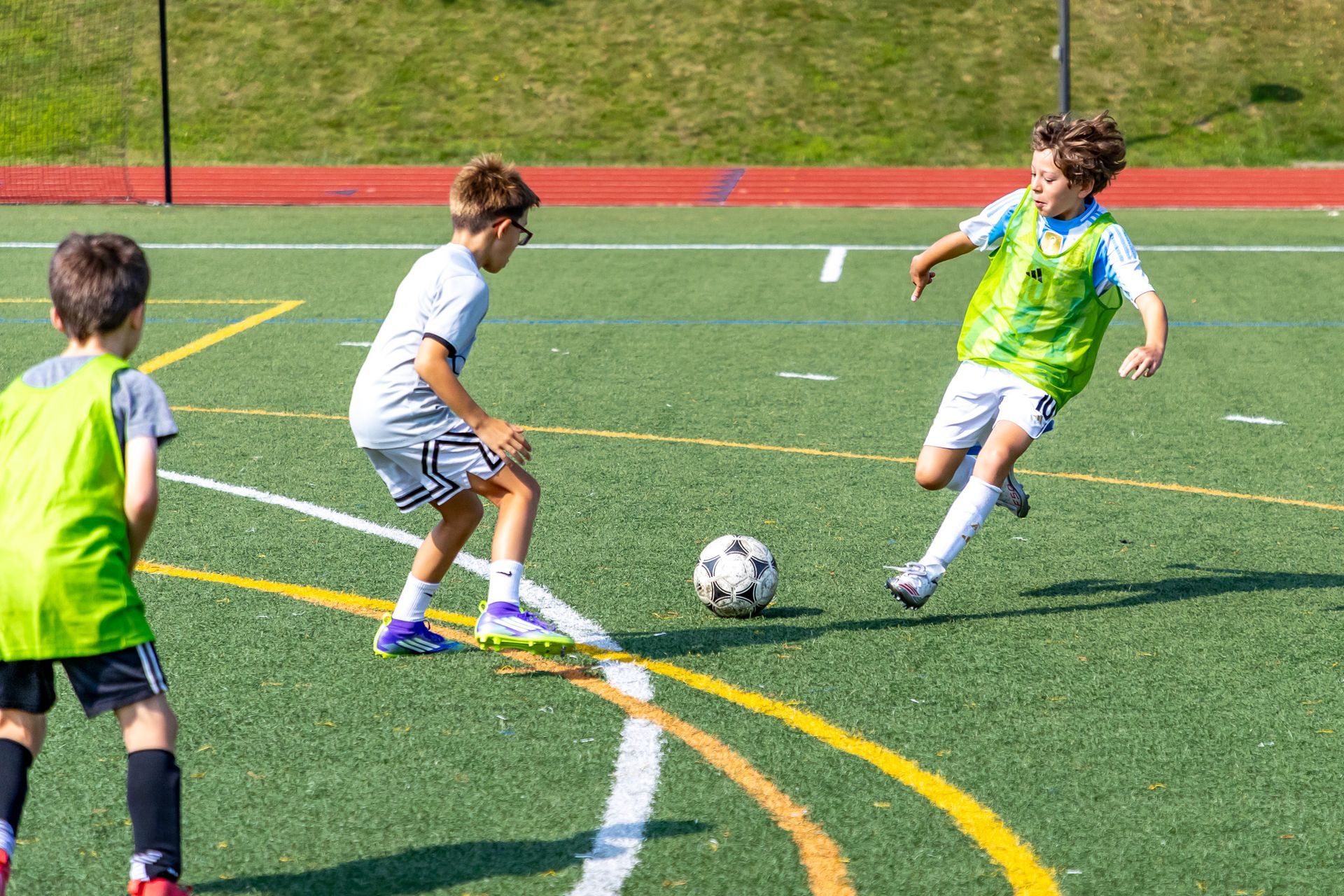 Three boys playing soccer on a green field. One boy in green vest kicks ball, two others watch.