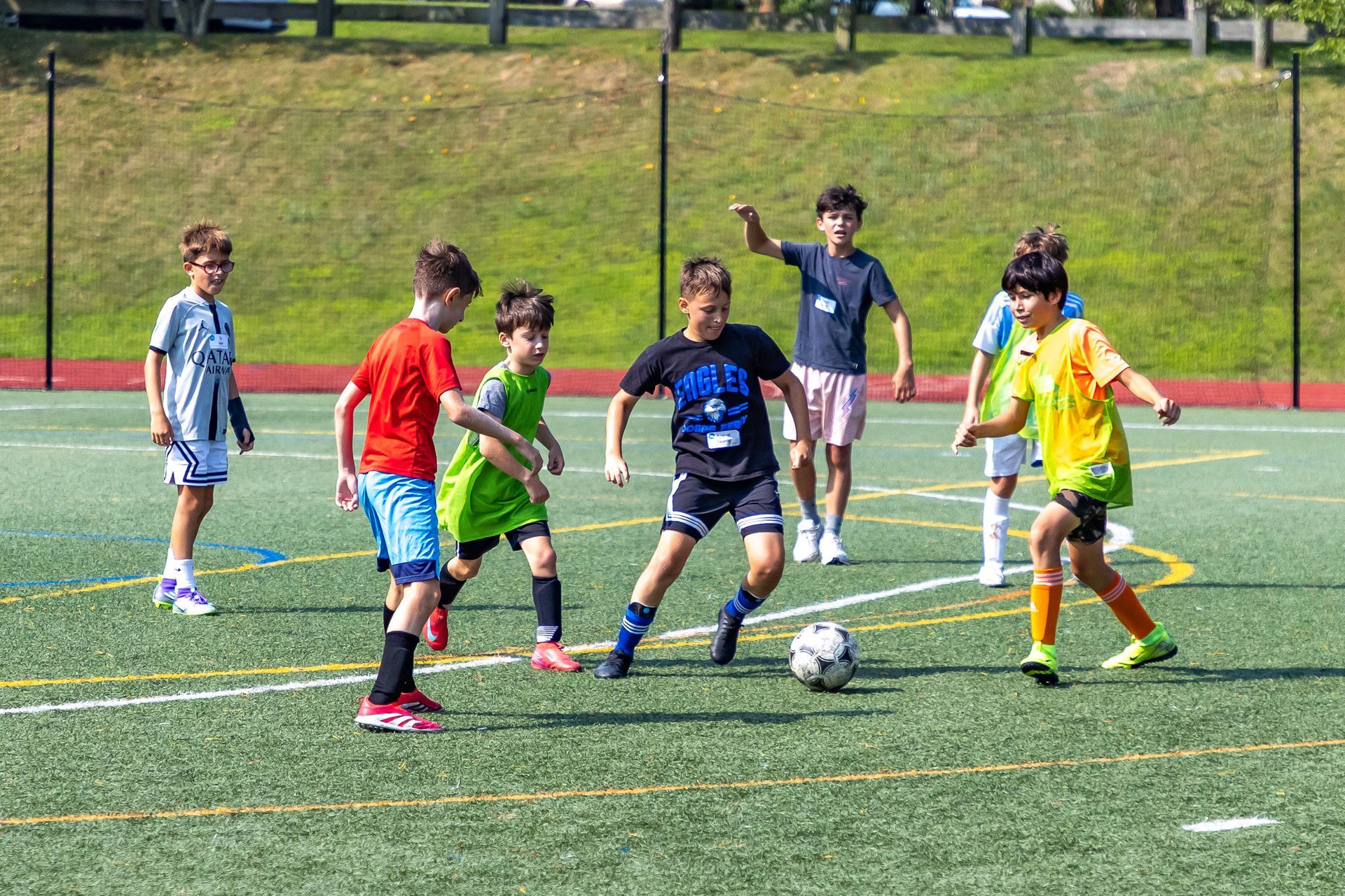 Kids playing soccer on a green field; boy in black shirt dribbles ball while others watch and move. Sunny day.