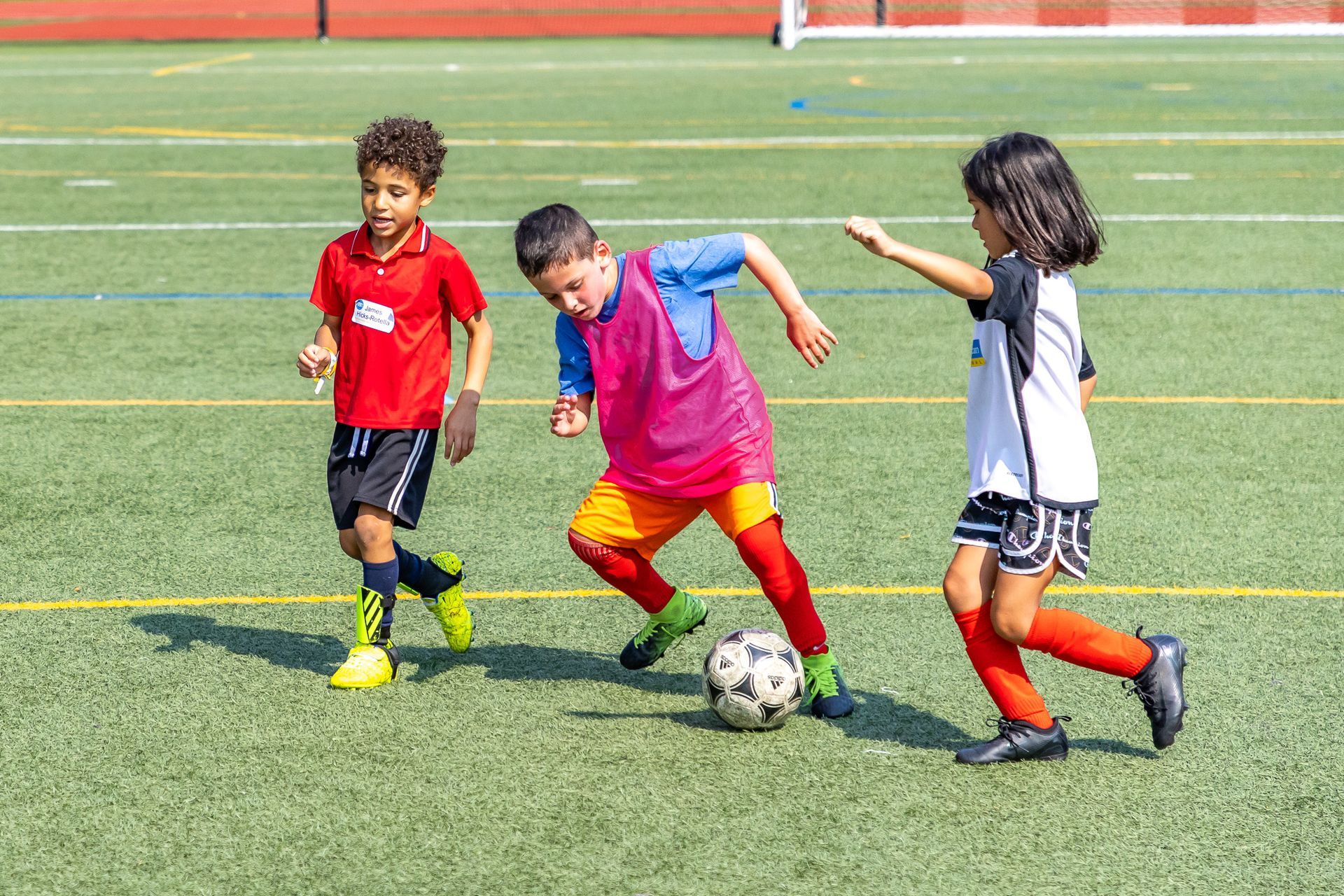 Three children playing soccer on a green field. One in red shirt dribbles the ball, others run alongside.