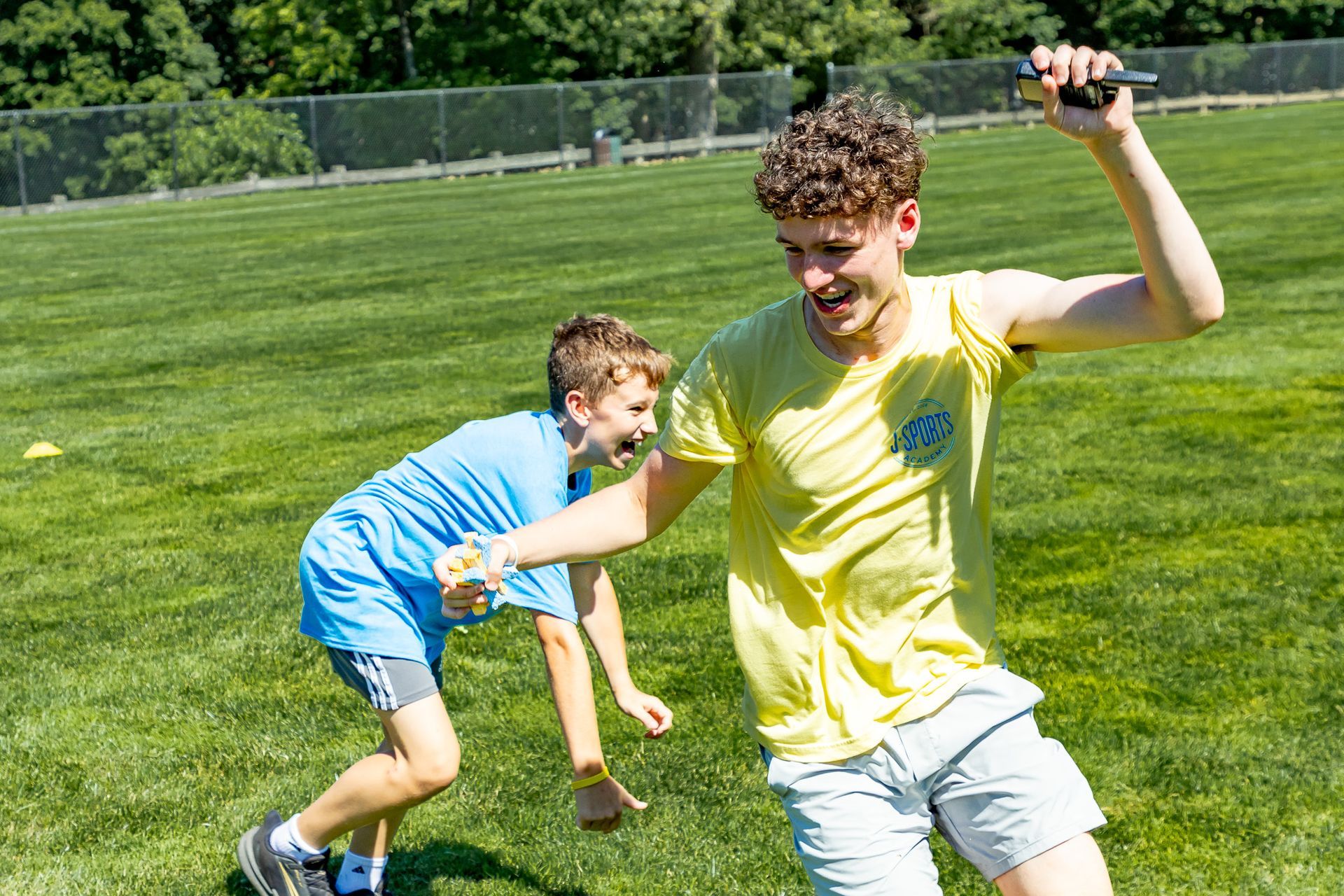 Two boys playing on a grassy field. One in blue shirt and shorts runs from boy in yellow, who raises a phone.