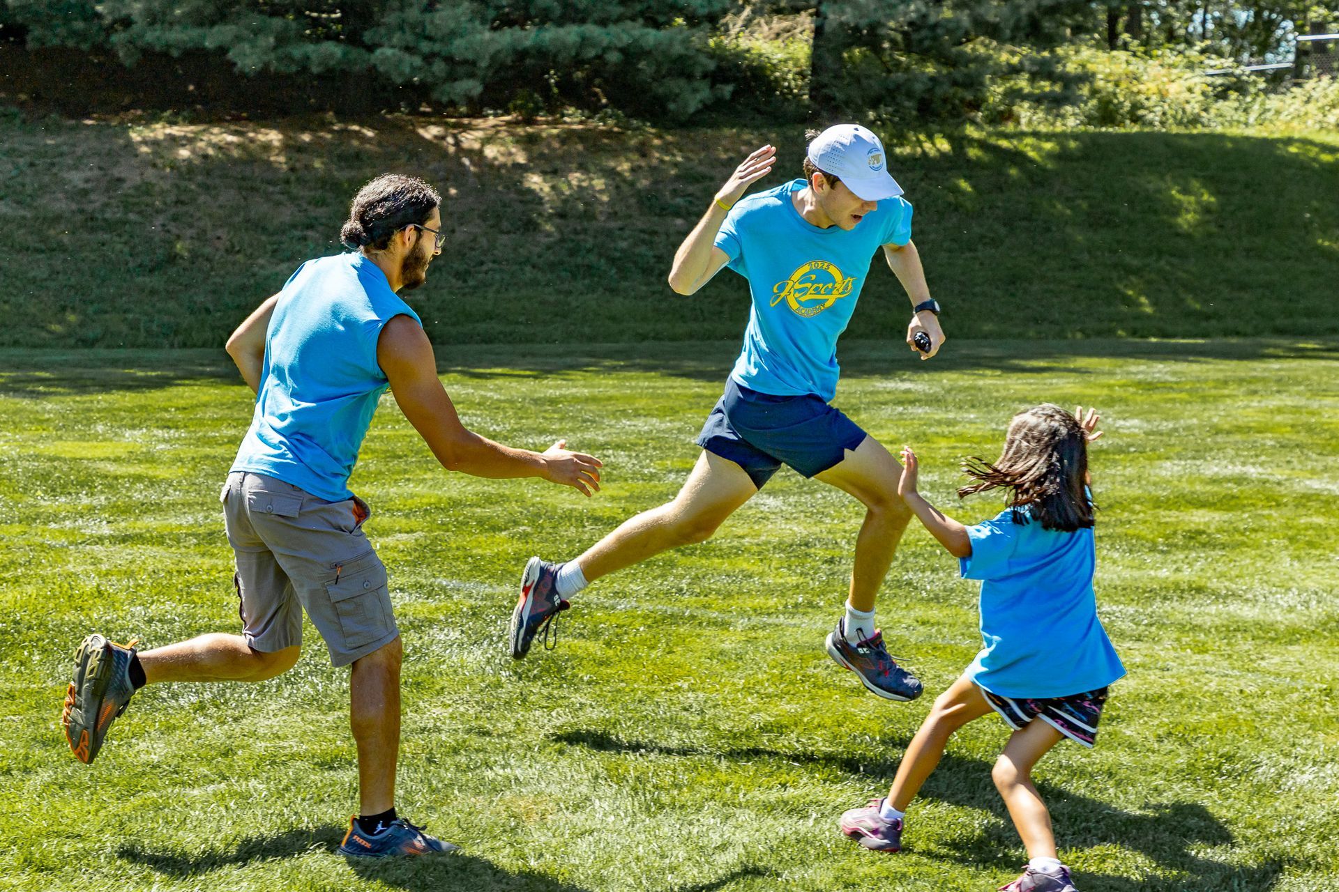 Three people playing tag in a grassy park: two men and a child. All wearing blue, one man jumping.