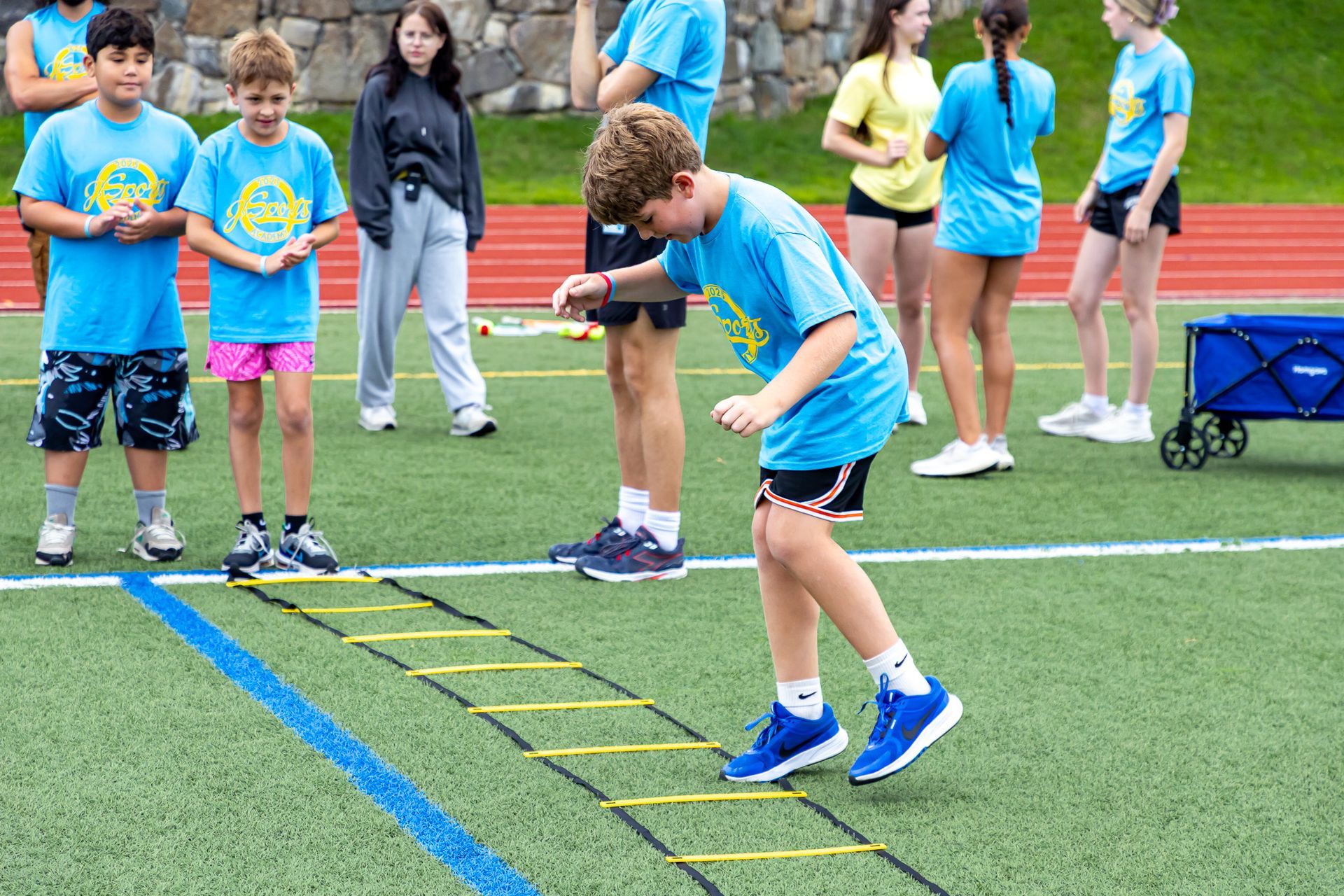 Children in blue shirts use an agility ladder on a track field.