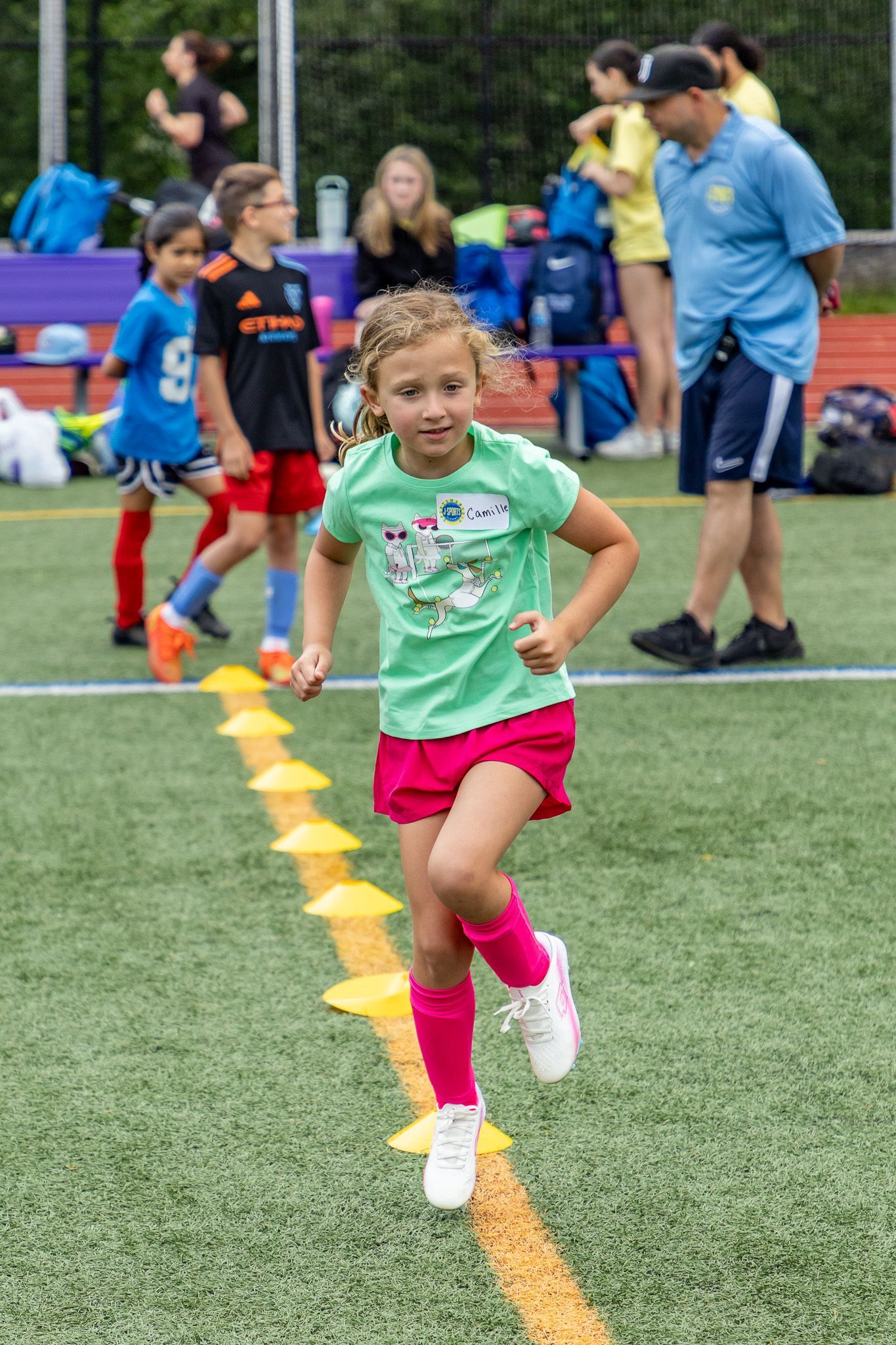 Young girl in pink shorts and socks runs a drill on a soccer field with orange cones.