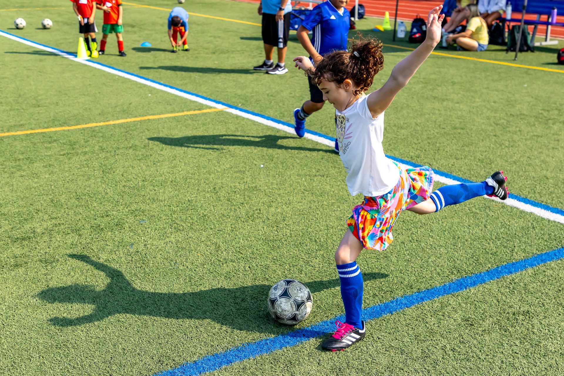 Girl in colorful shorts kicks a soccer ball on a green field, arms raised. Other players and adults in the background.