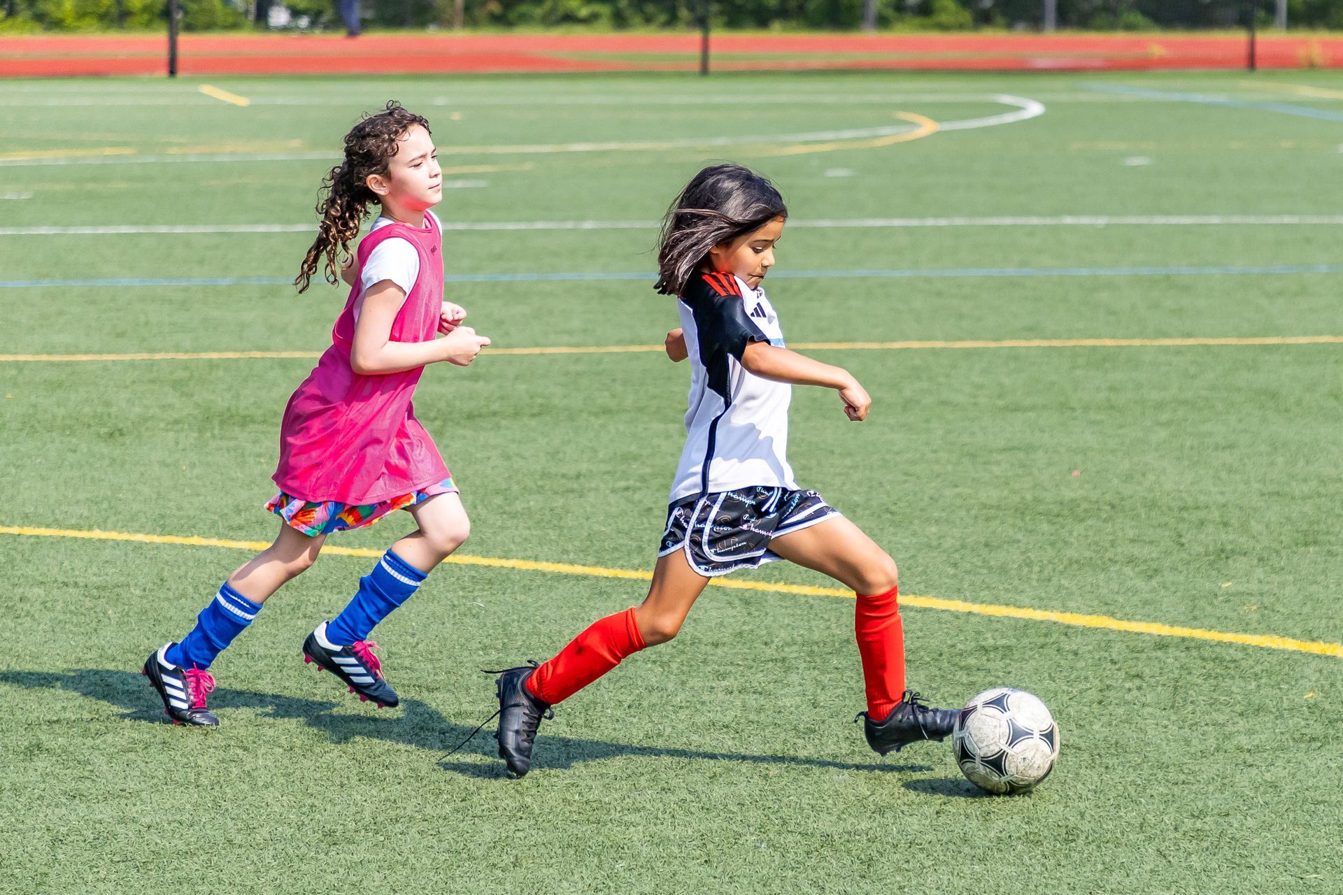 Two girls playing soccer on a green field; one kicking the ball, the other running behind.