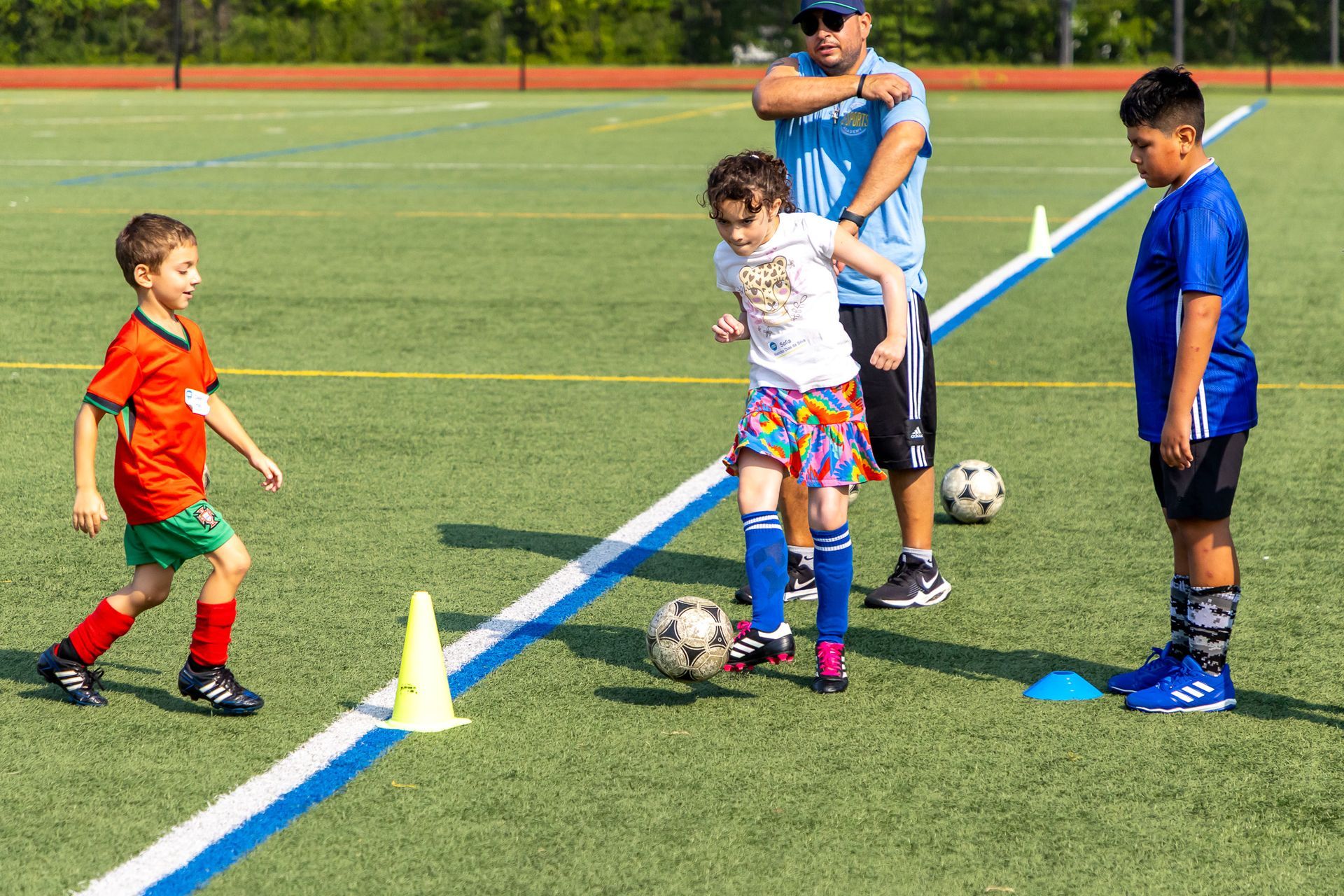 Youth soccer training on a green field: kids dribbling, coach watching, cones mark a path.