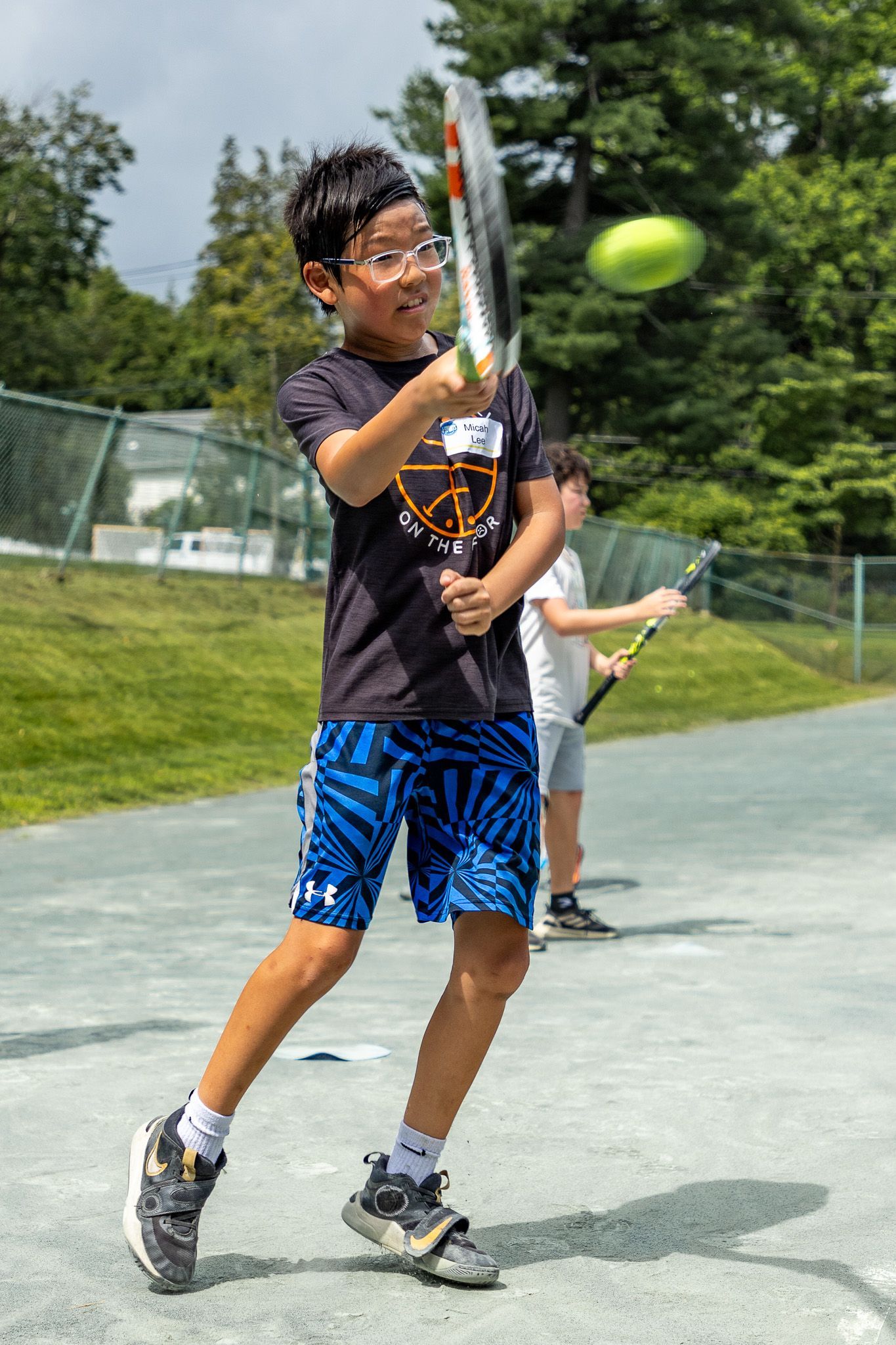 Boy in glasses swings a tennis racket on a court, hitting a yellow ball. Another player in background.