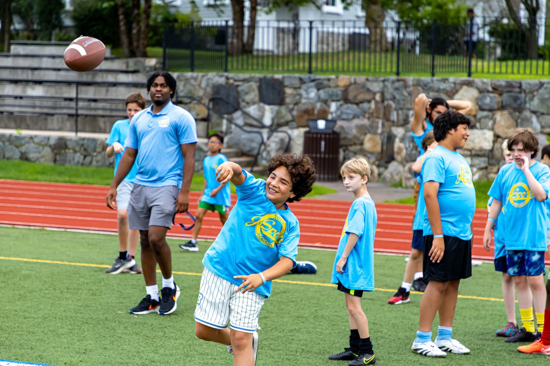 Kids and counselor playing football on a field; one boy throws, others watch. Blue shirts, sunny day.