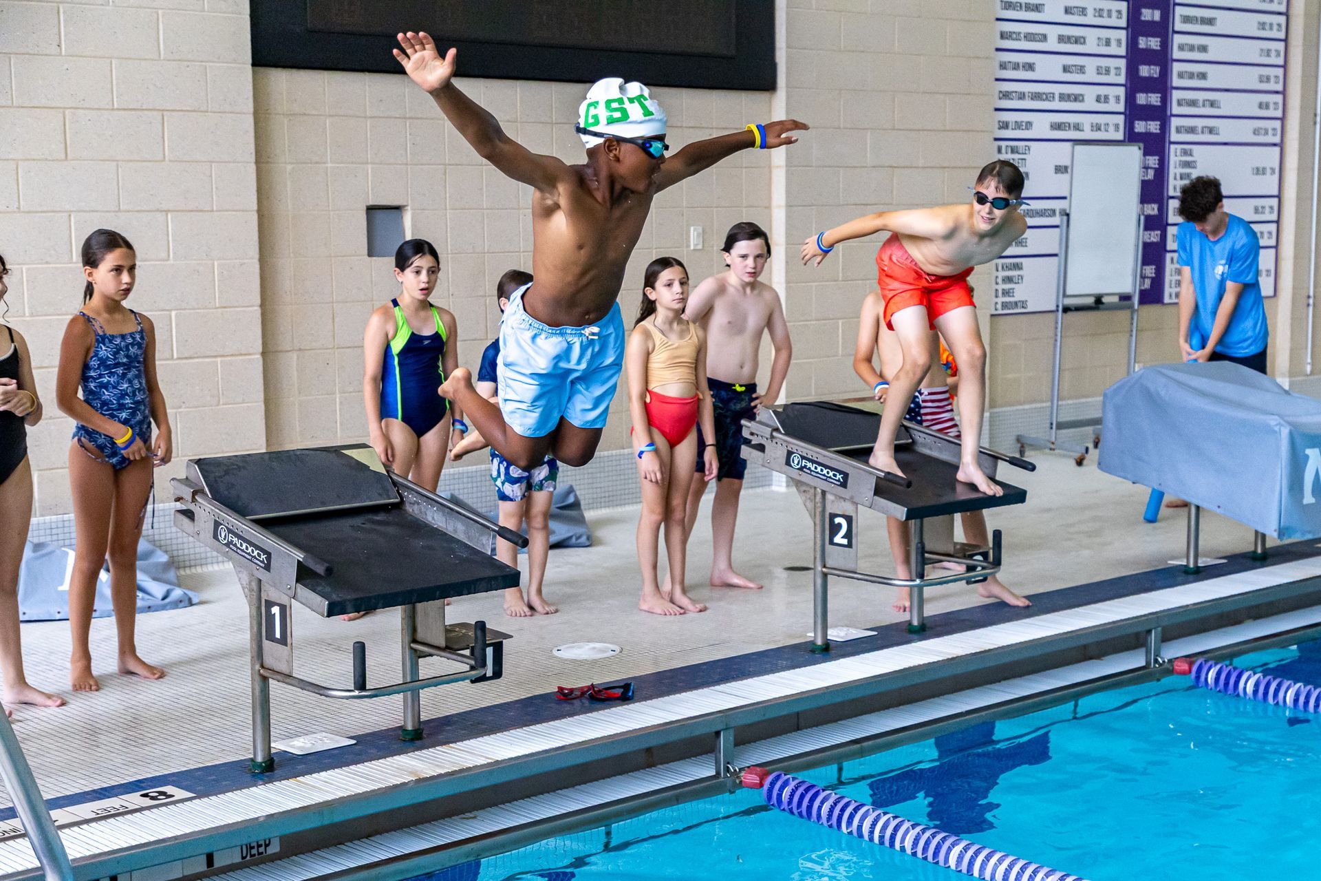Two swimmers leap from starting blocks in a pool, arms raised. Spectators watch.