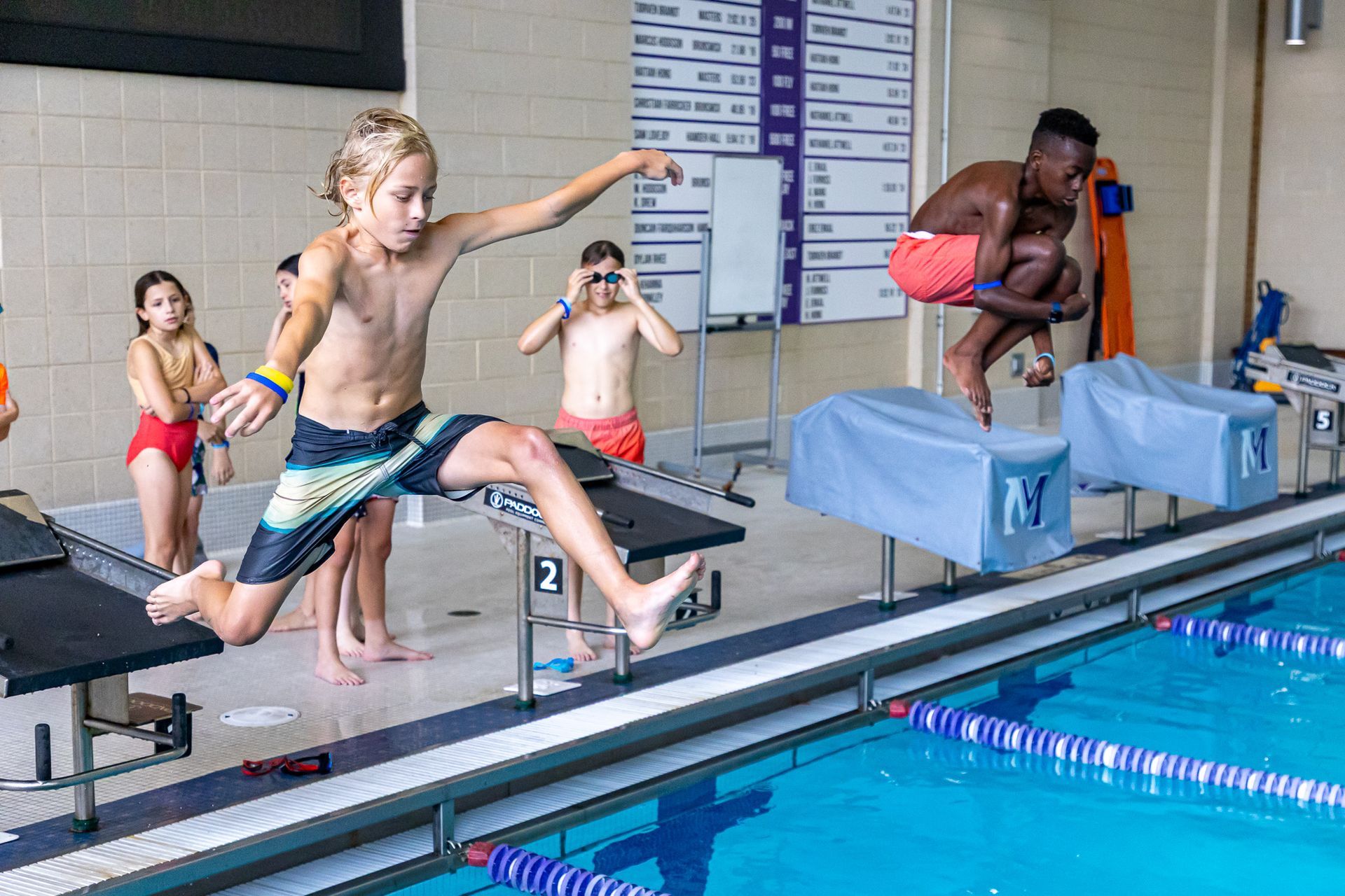 Children jumping into a pool from starting blocks; boy in black and teal swim trunks leads.