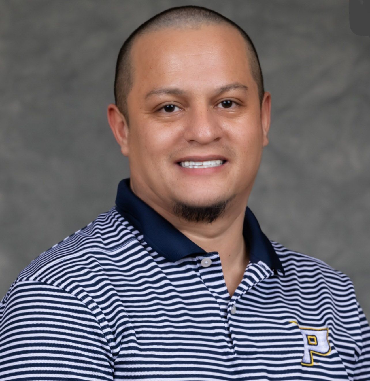A smiling man with a shaved head and goatee wearing a striped polo shirt with a 