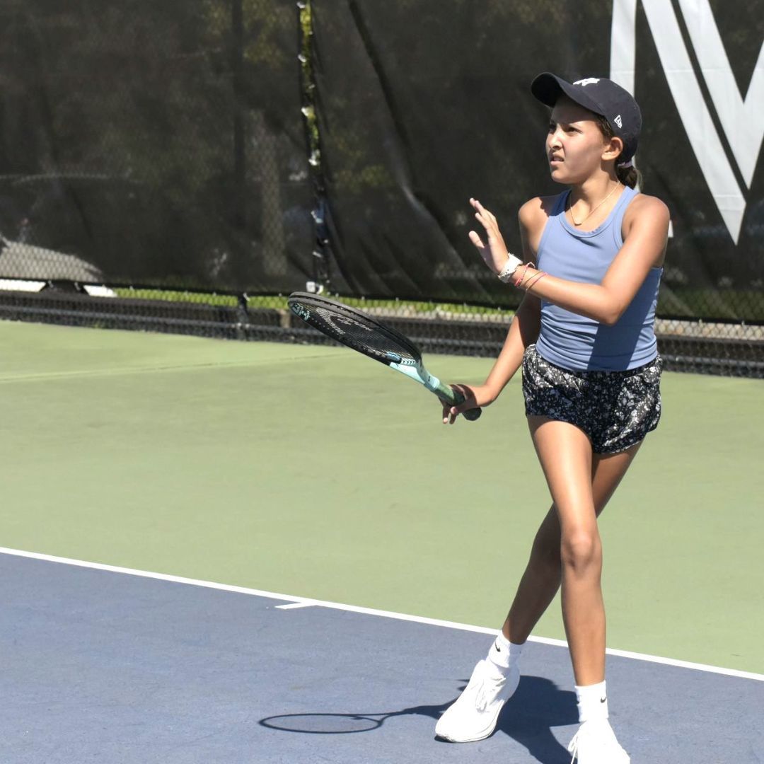 Young tennis player in blue tank top and black shorts swings racket on a blue and green court.