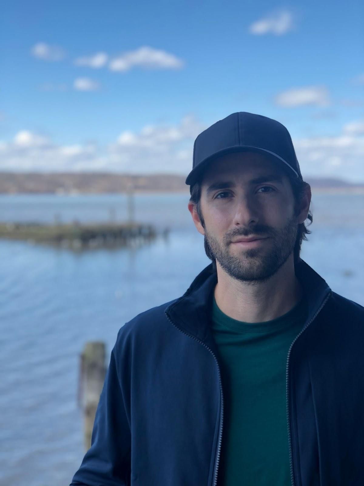 A man wearing a hat and a jacket stands in front of a body of water
