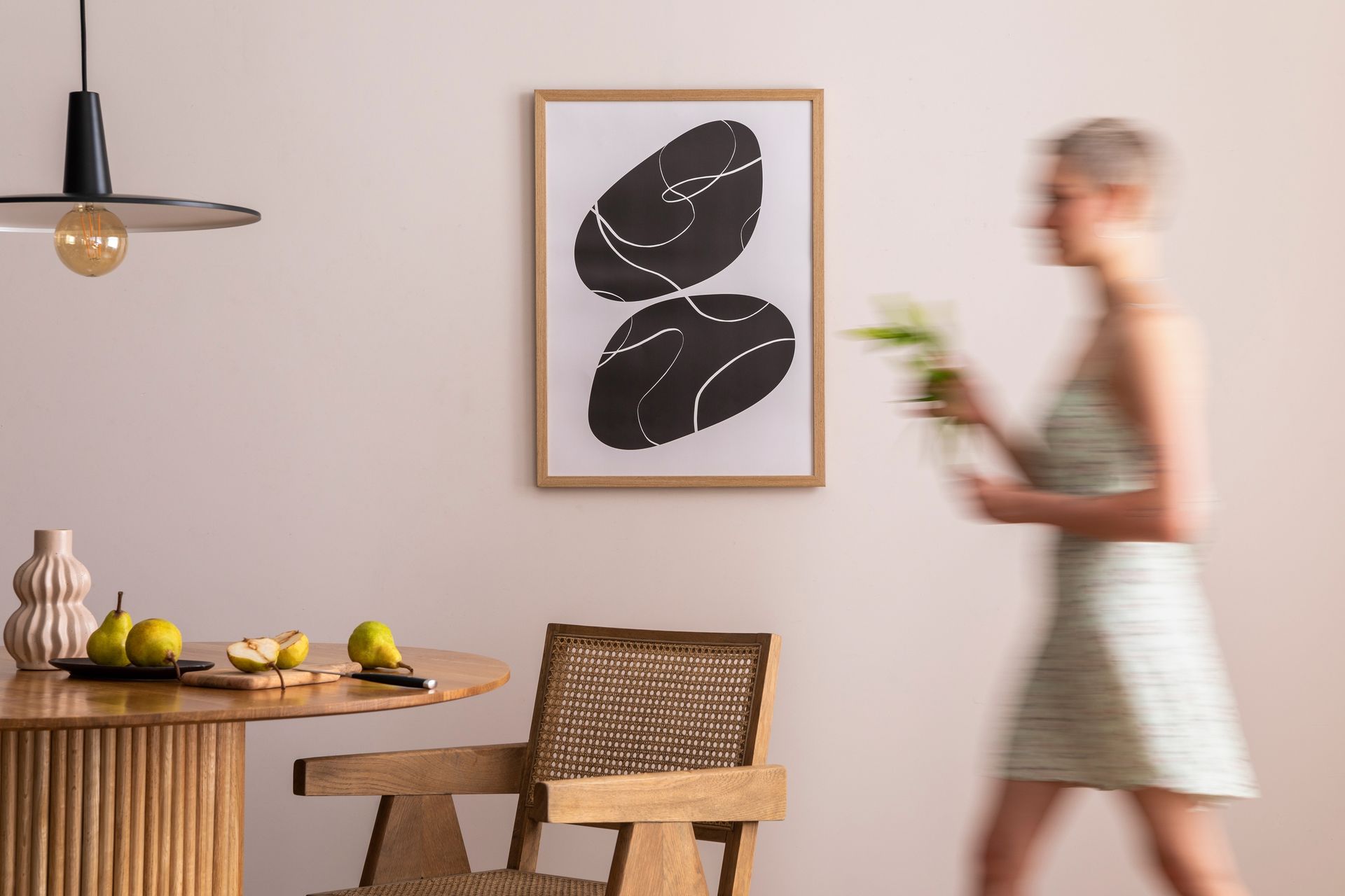 Modern dining room with wooden table, leaf wall art, and a blurred person walking past holding flowers