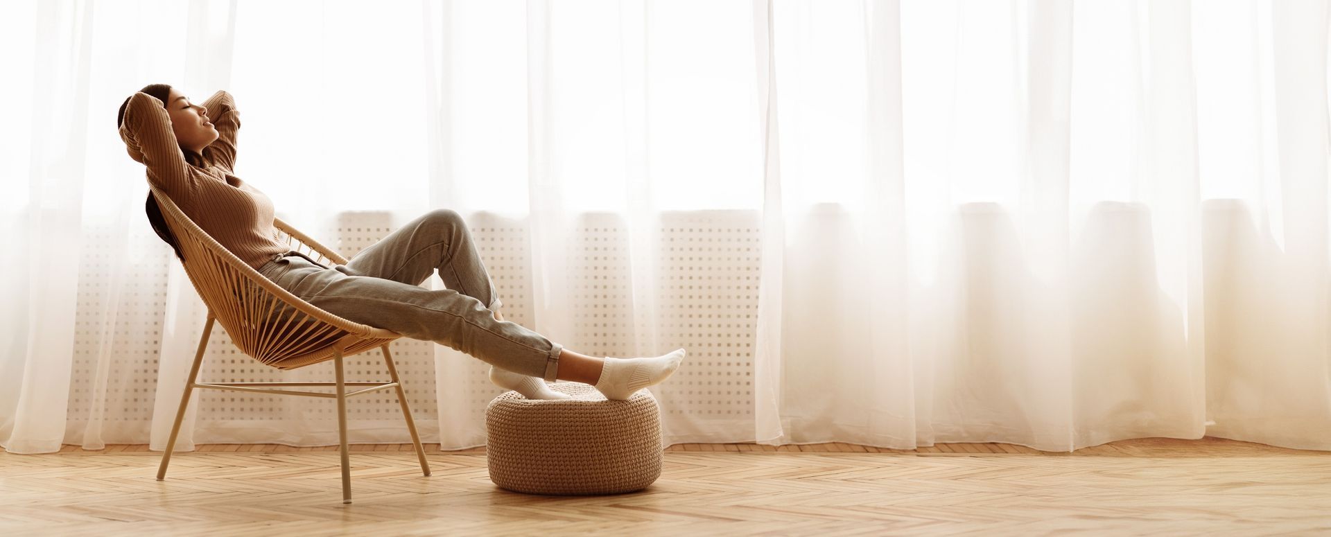 Person reclining in a wicker chair by sheer curtains, feet on a pouf in a sunlit room.