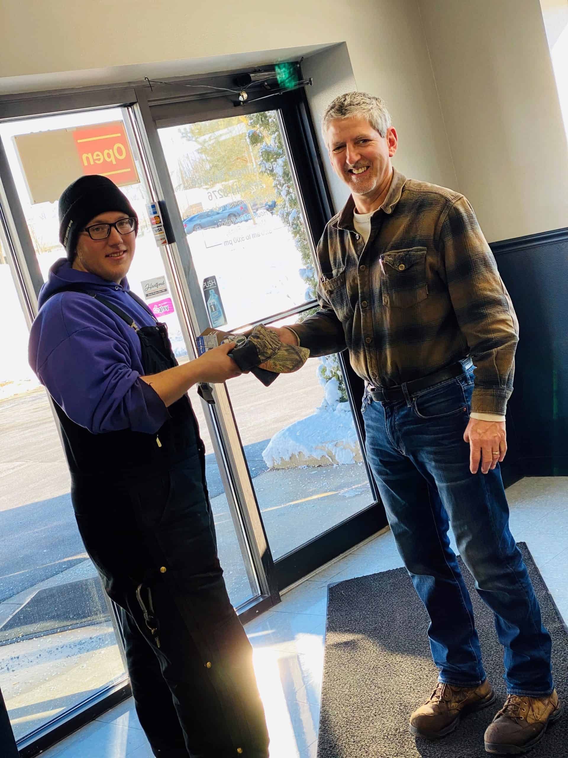 Two men shaking hands in front of a glass door. One wears a purple hoodie and overalls, the other a plaid shirt and jeans. | Joe's Slinger Service